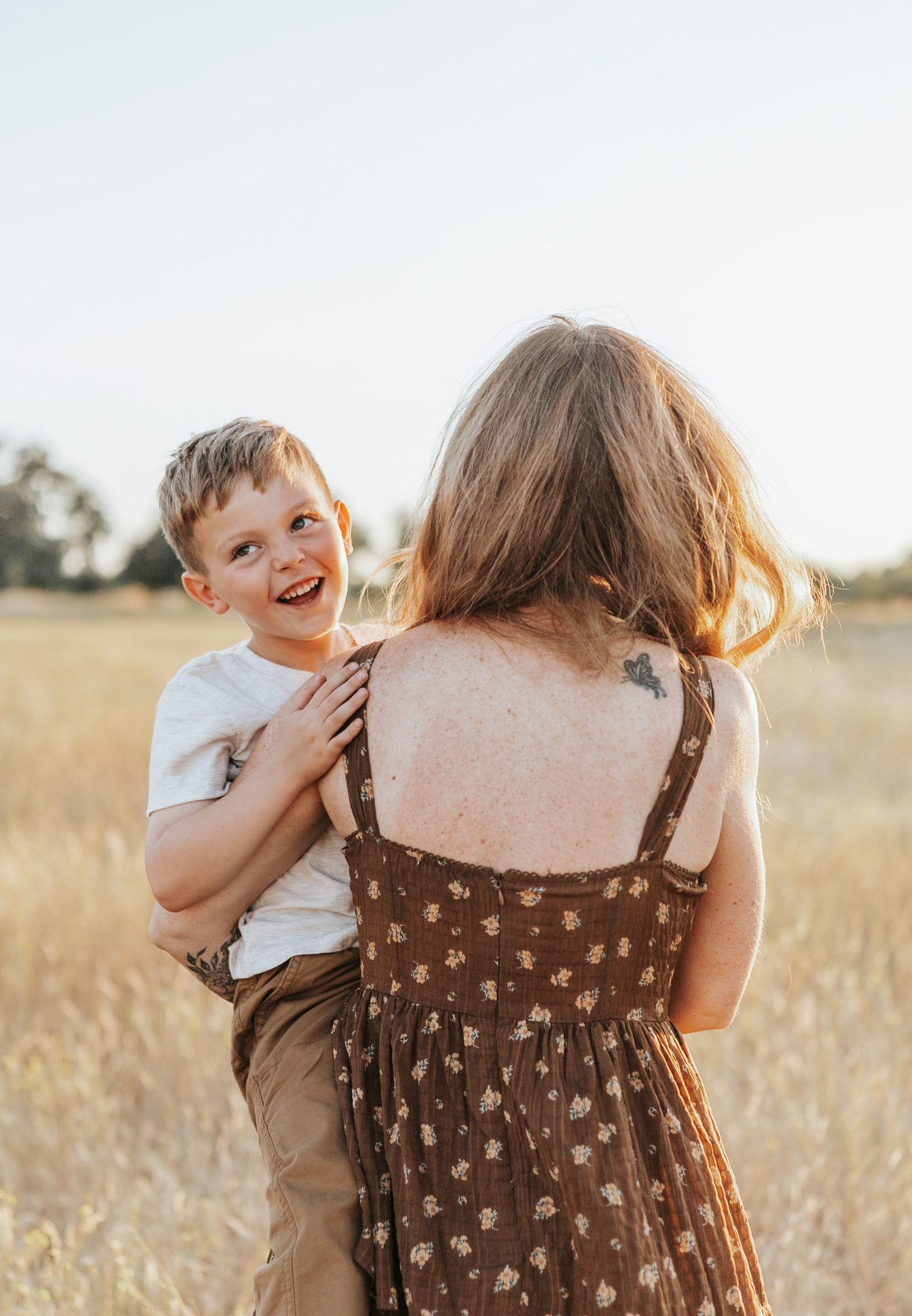 A joyful child plays and laughs with someone in a floral dress against a backdrop of golden grass at sunset.