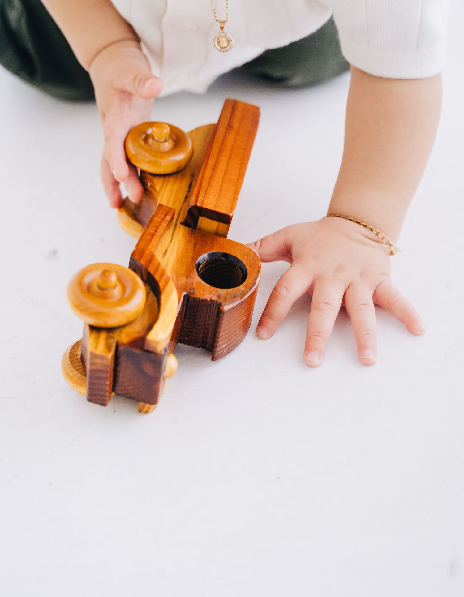 Child playing with a handcrafted wooden toy train on a white surface, wearing a gold bracelet.