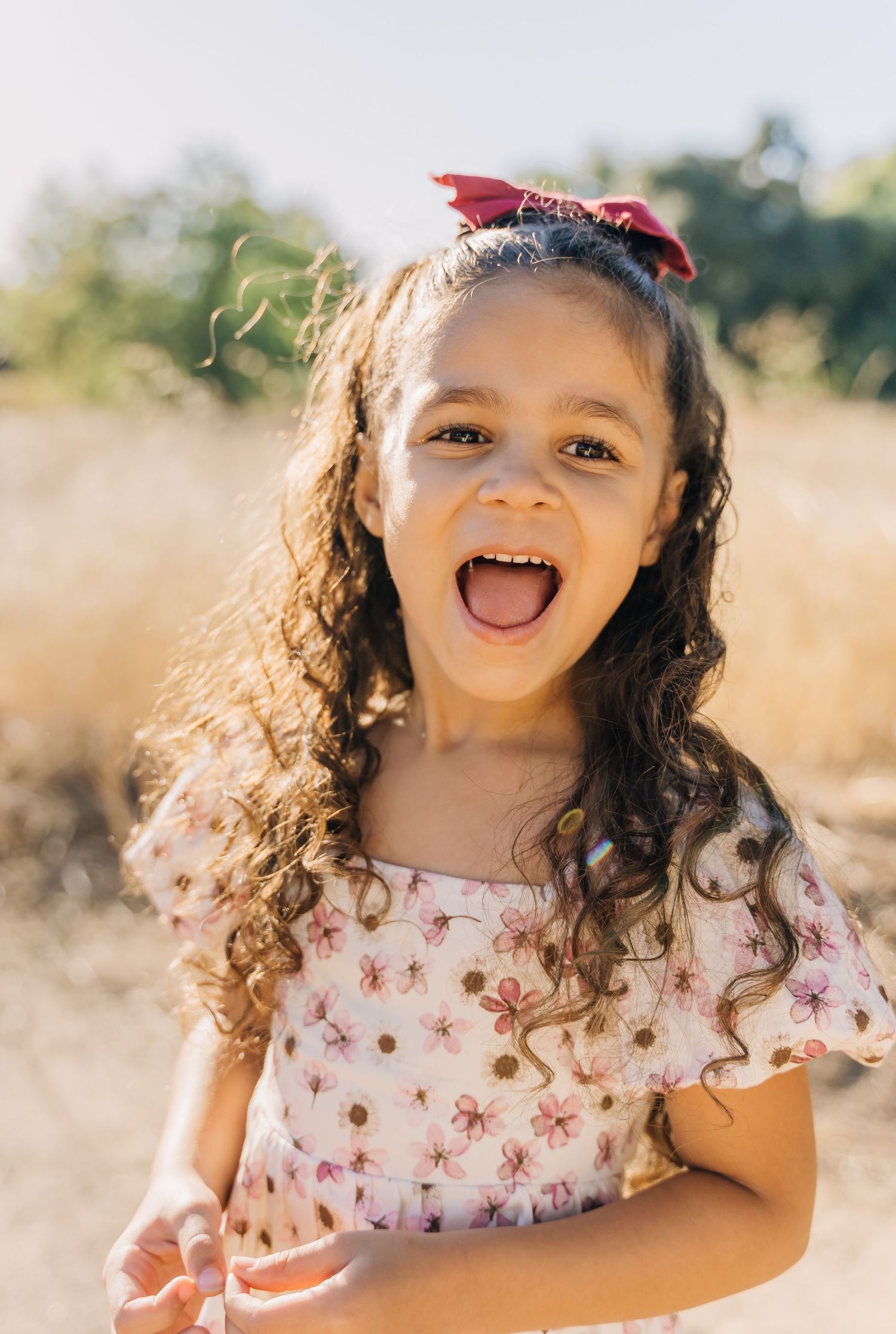A child wearing a floral dress and red hair bow smiles joyfully in a sunny outdoor field.