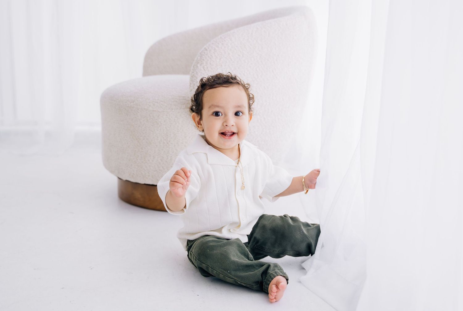Smiling baby in white shirt and green pants sitting on floor next to a modern white chair in bright room.