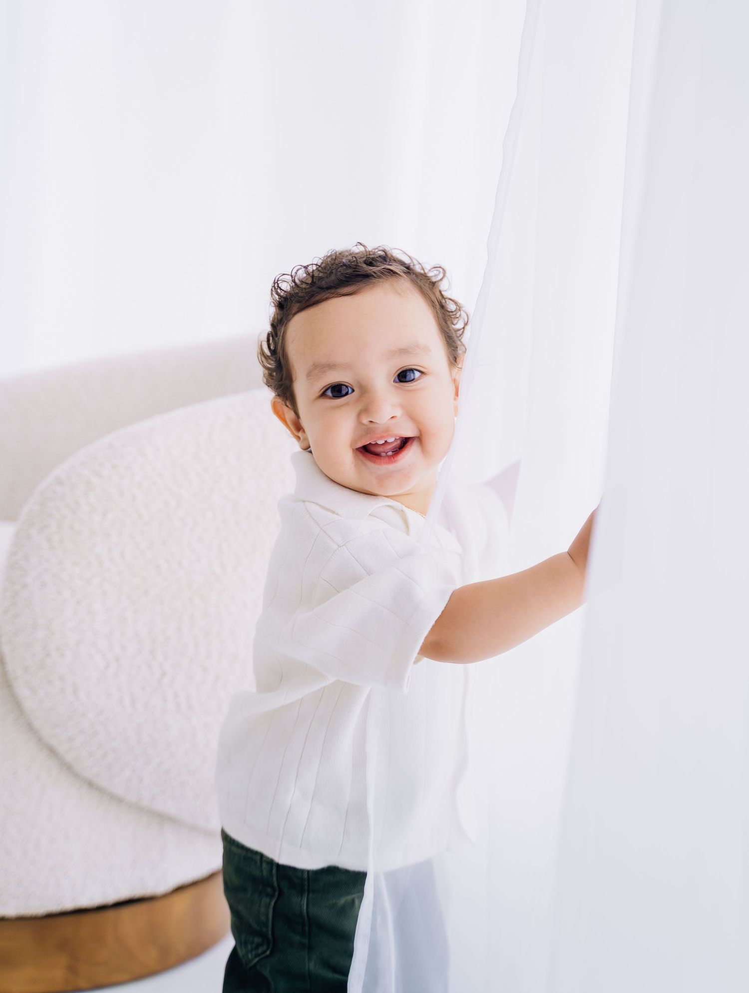 Smiling toddler in white shirt and green pants holding a sheer curtain in a bright white studio.