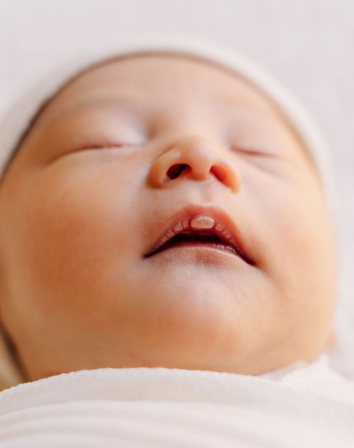 Close-up of a peaceful sleeping newborn baby wearing a white hat, swaddled in a soft white blanket.