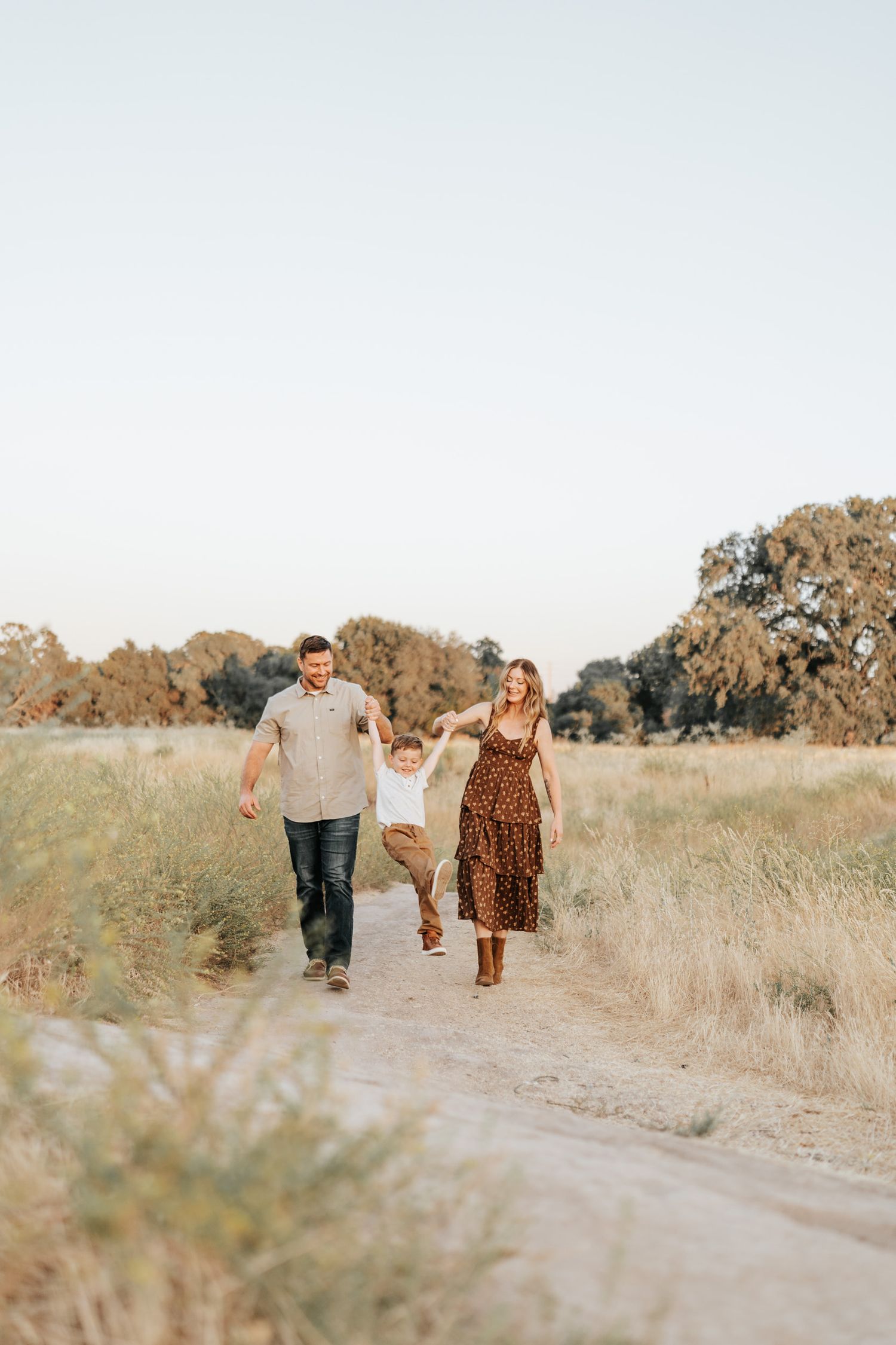 Family walks together holding hands on a dirt path through a golden autumn field at sunset.