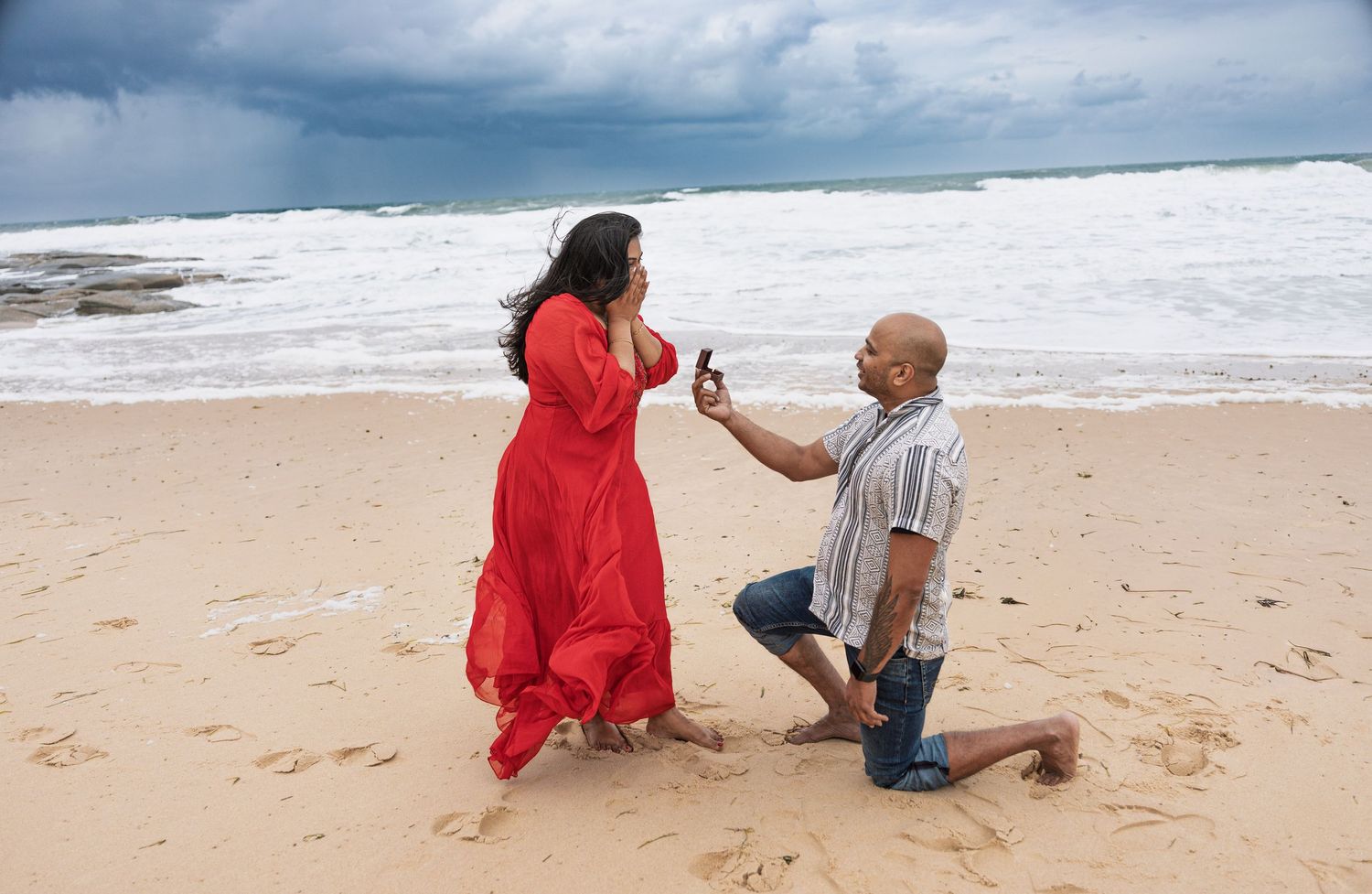 A seaside proposal unfolds with dramatic ocean waves and cloudy skies.