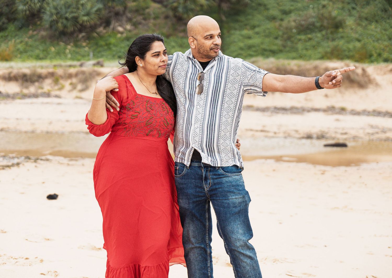 Couple in red dress and striped shirt pointing while standing on beach.