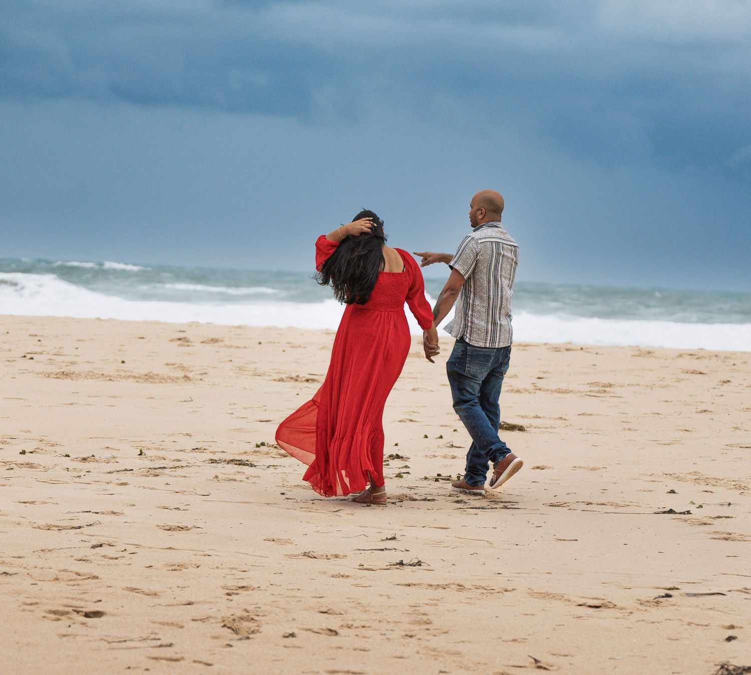 Couple walking hand in hand on empty beach with moody sky and ocean waves.