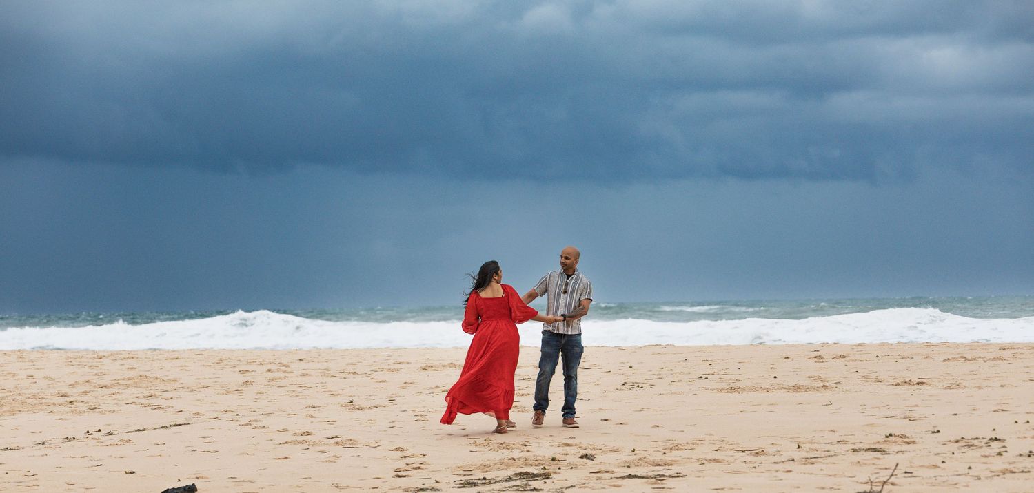 Two people strolling along sandy beach with dramatic cloudy sky and ocean waves.