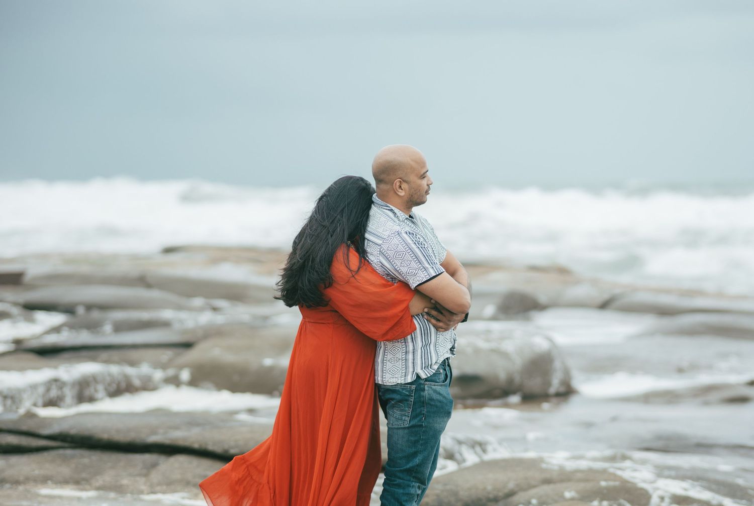 A couple shares tender moments at a rocky beach location with waves crashing in the background.