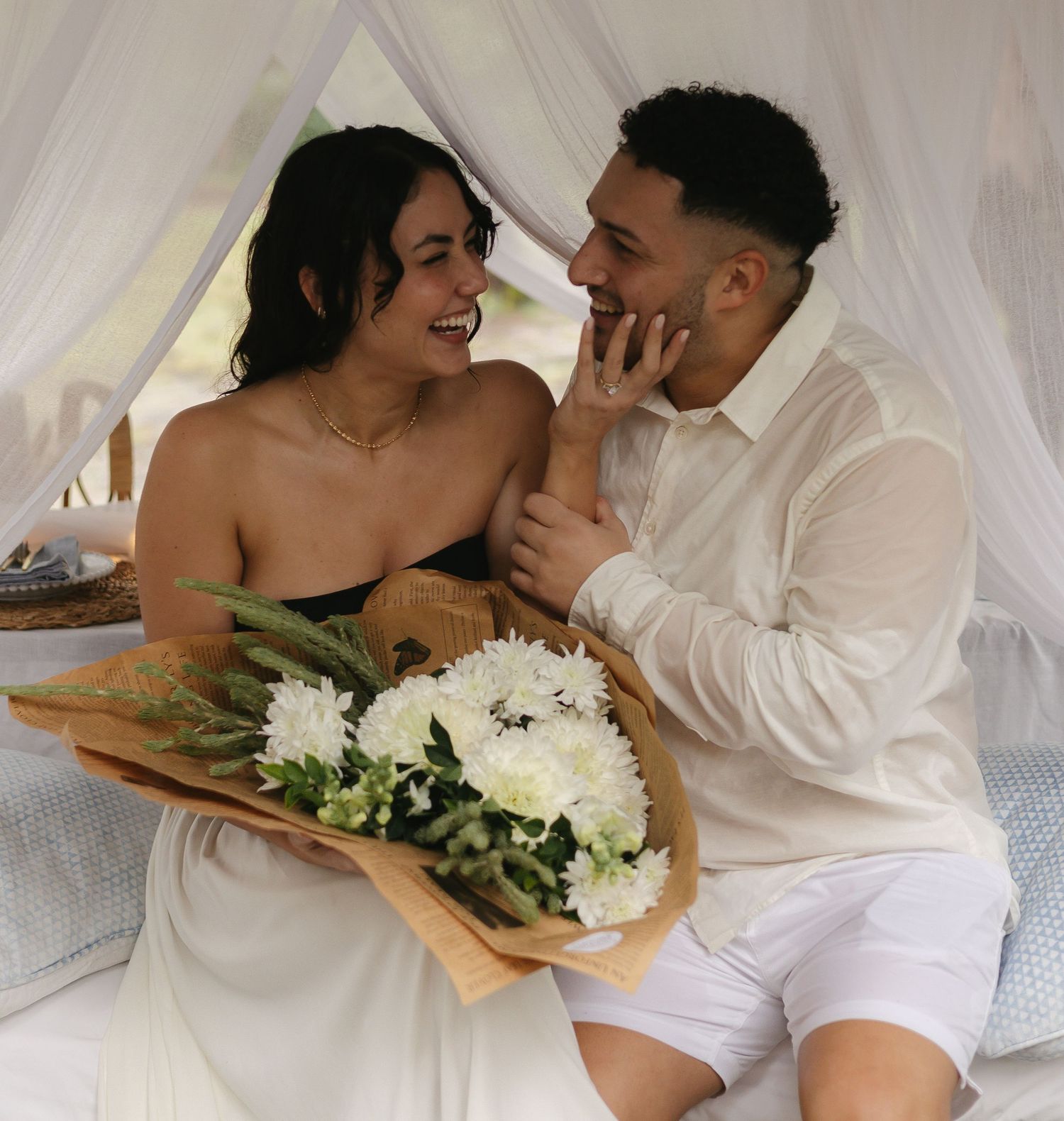 Romantic couple with white floral bouquet under flowing fabric canopy.