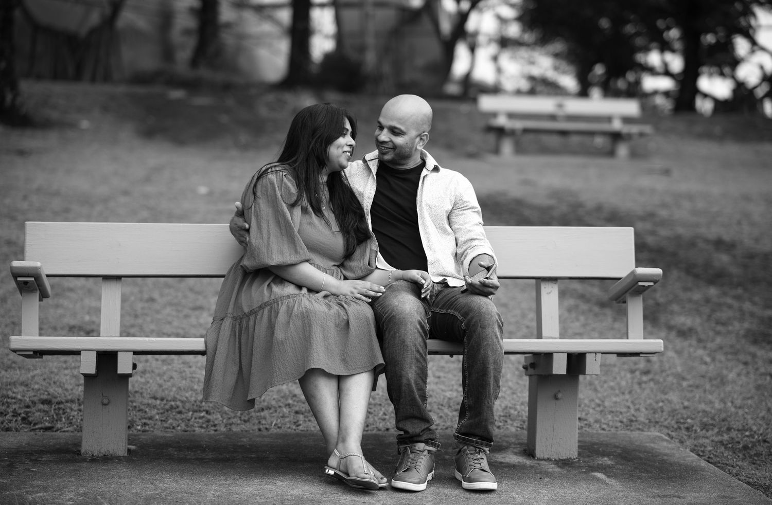 A couple sits close together on white benches in a tranquil park setting.