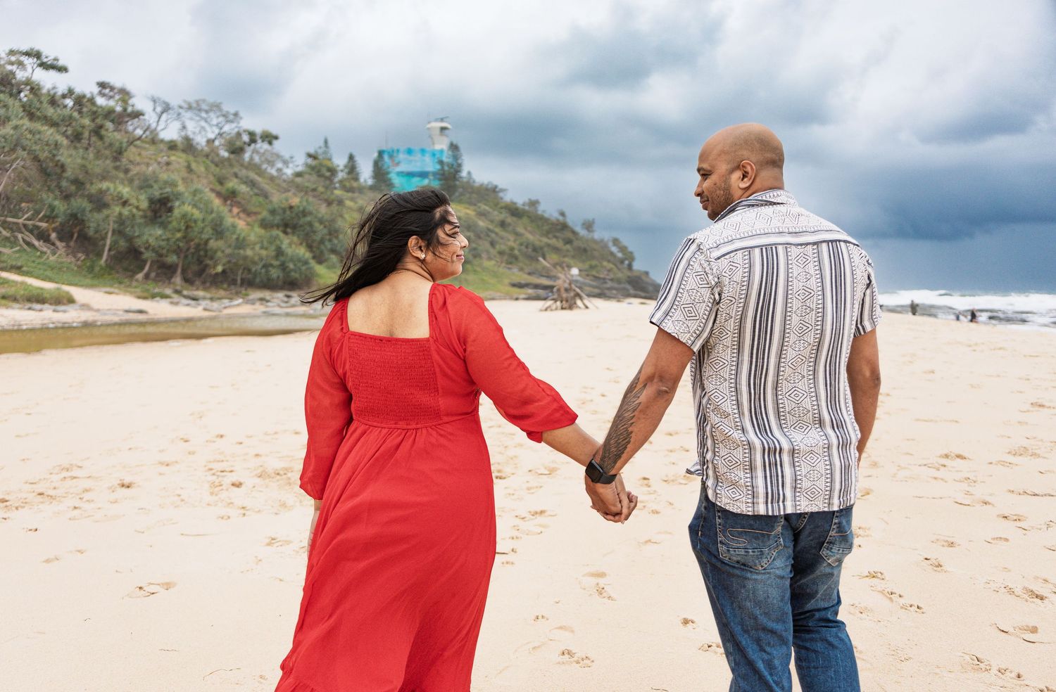 Couple holding hands while walking on scenic coastal beach under cloudy sky.