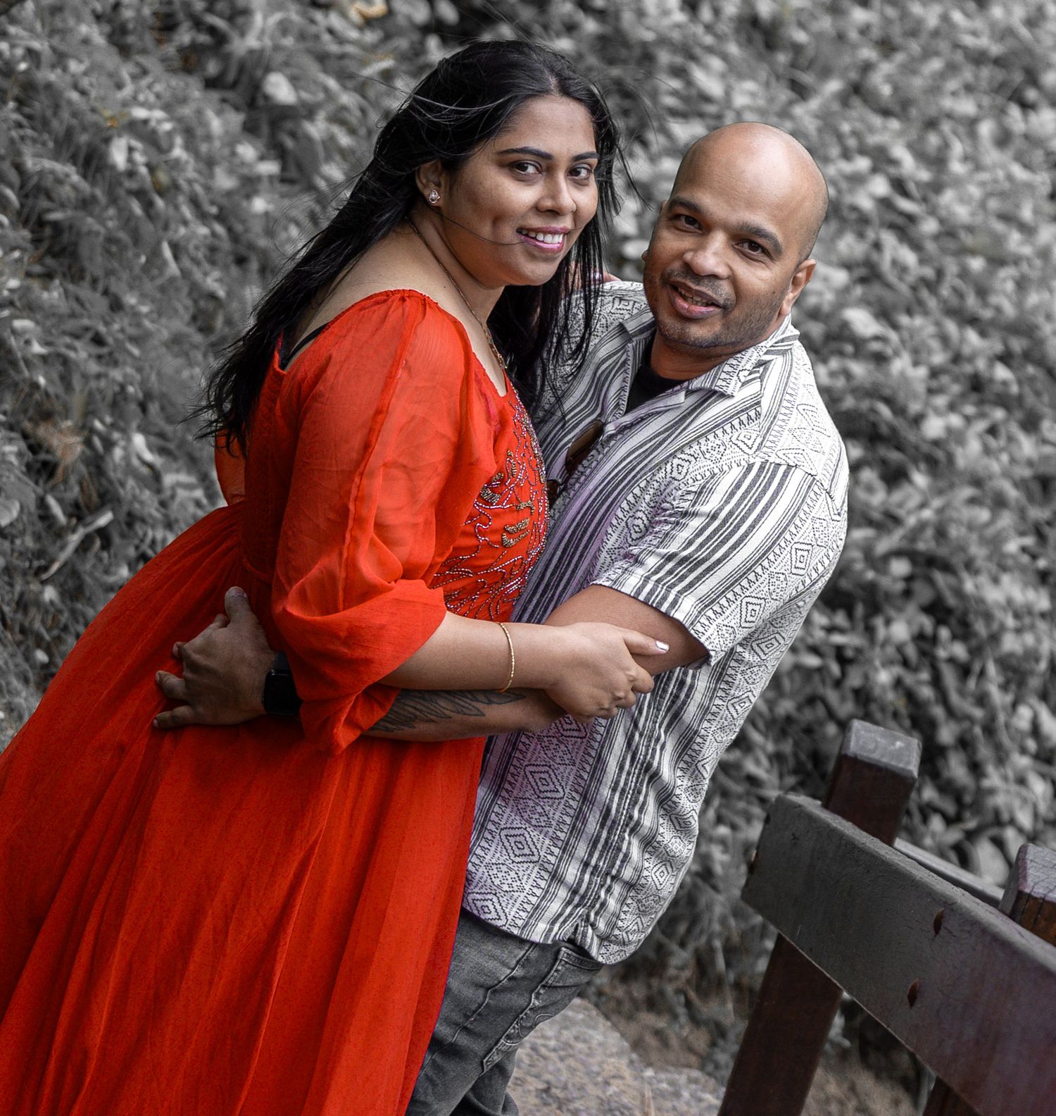 A couple embraces outdoors wearing red dress and striped shirt against a monochromatic background.