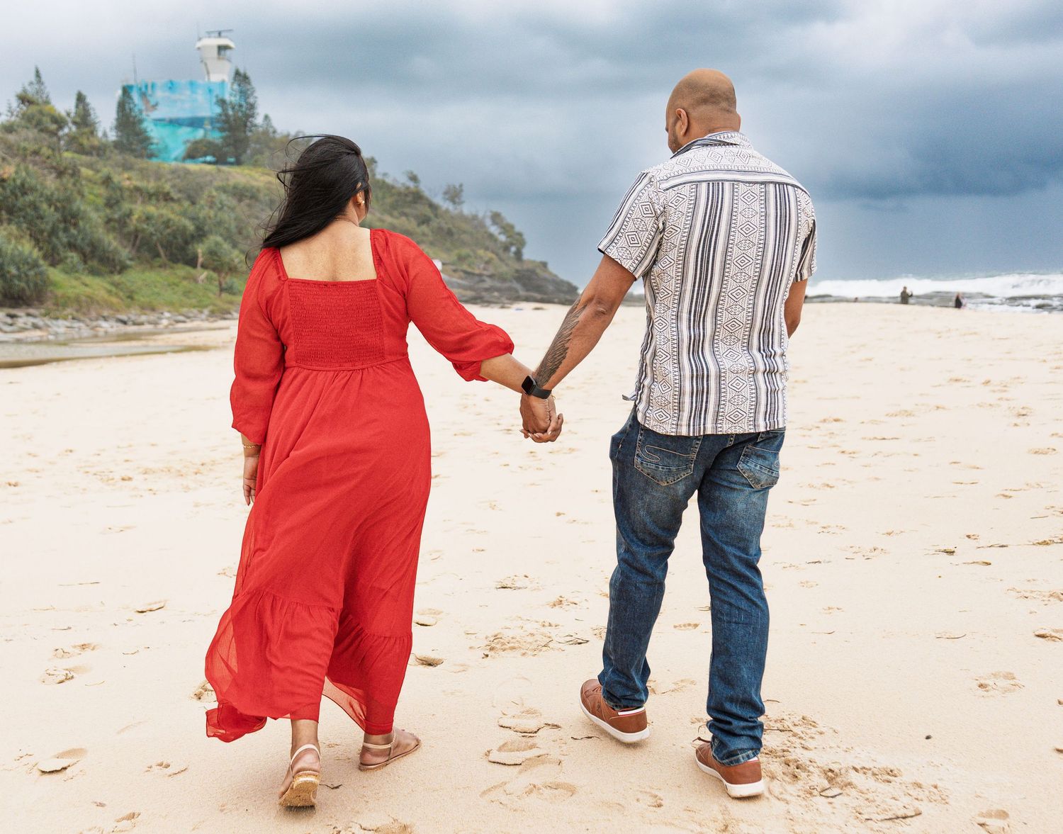 Two people on beach wearing red dress and striped shirt walking away from camera.