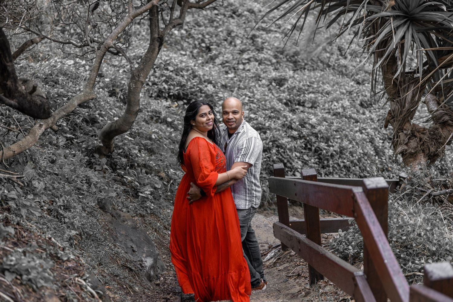 Couple poses on nature trail with woman in flowing red dress and man in striped shirt.