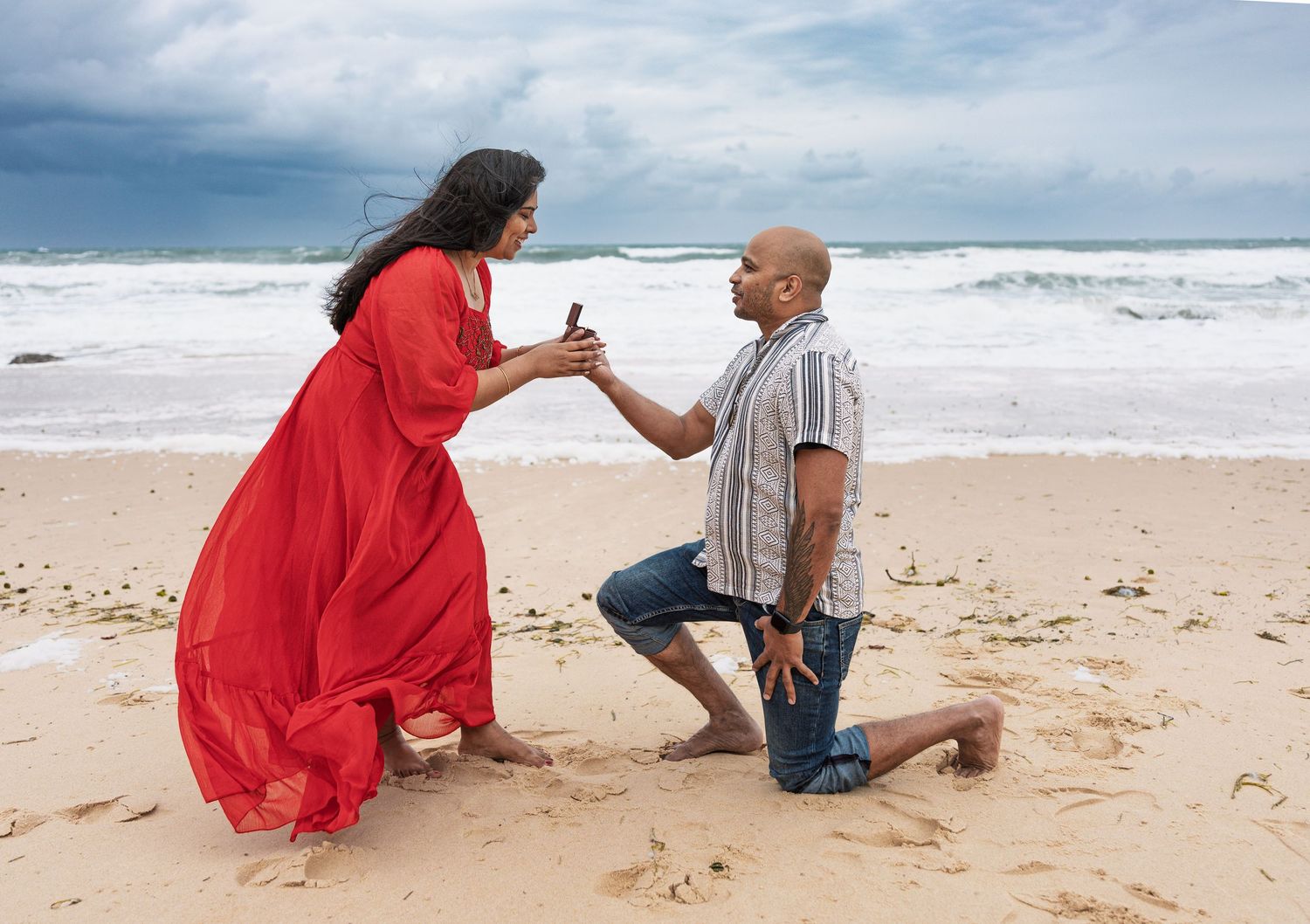 A couple embraces on a scenic beach with crashing waves and cloudy skies.