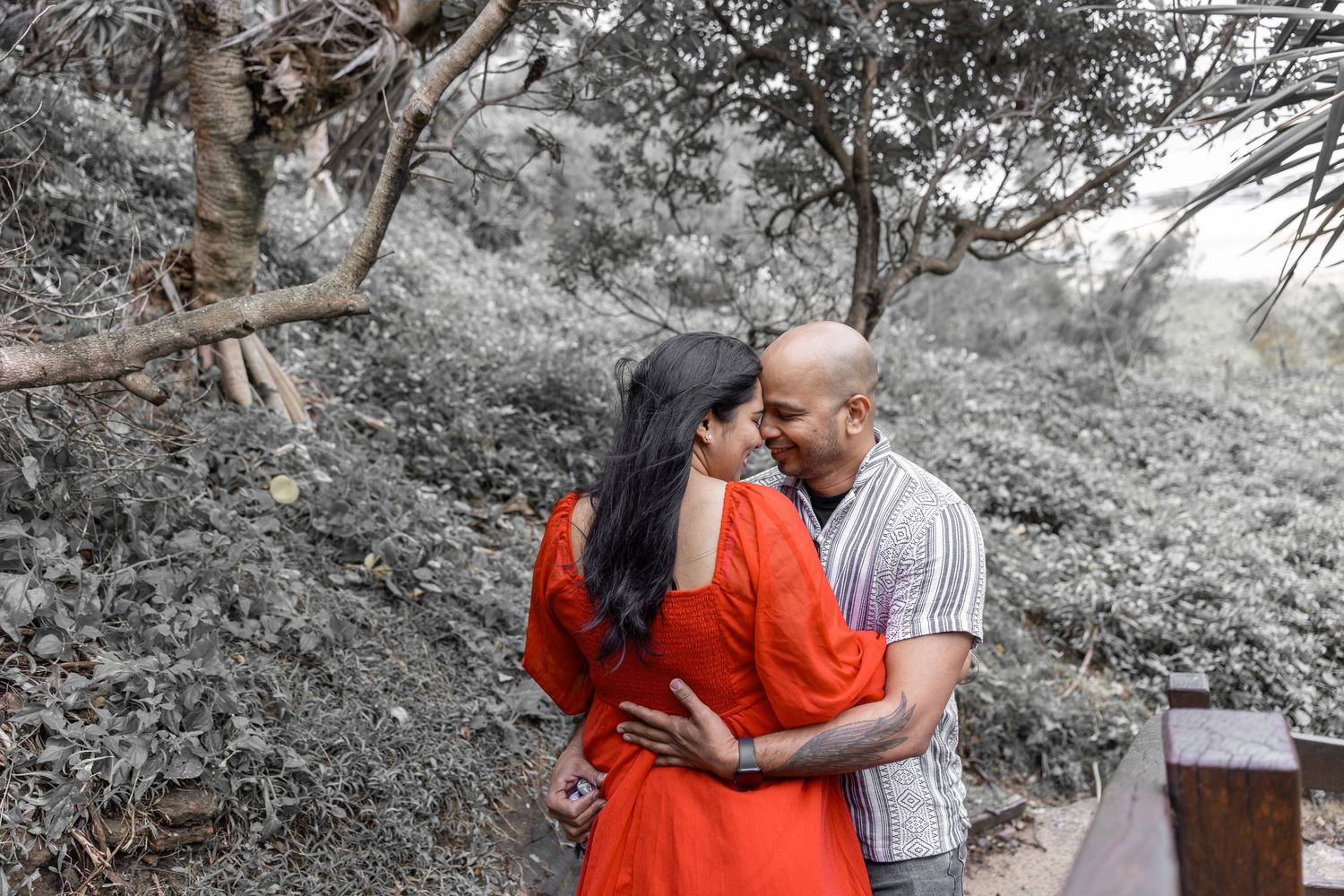 Tender moment between couple in red dress and striped shirt against desaturated nature backdrop.