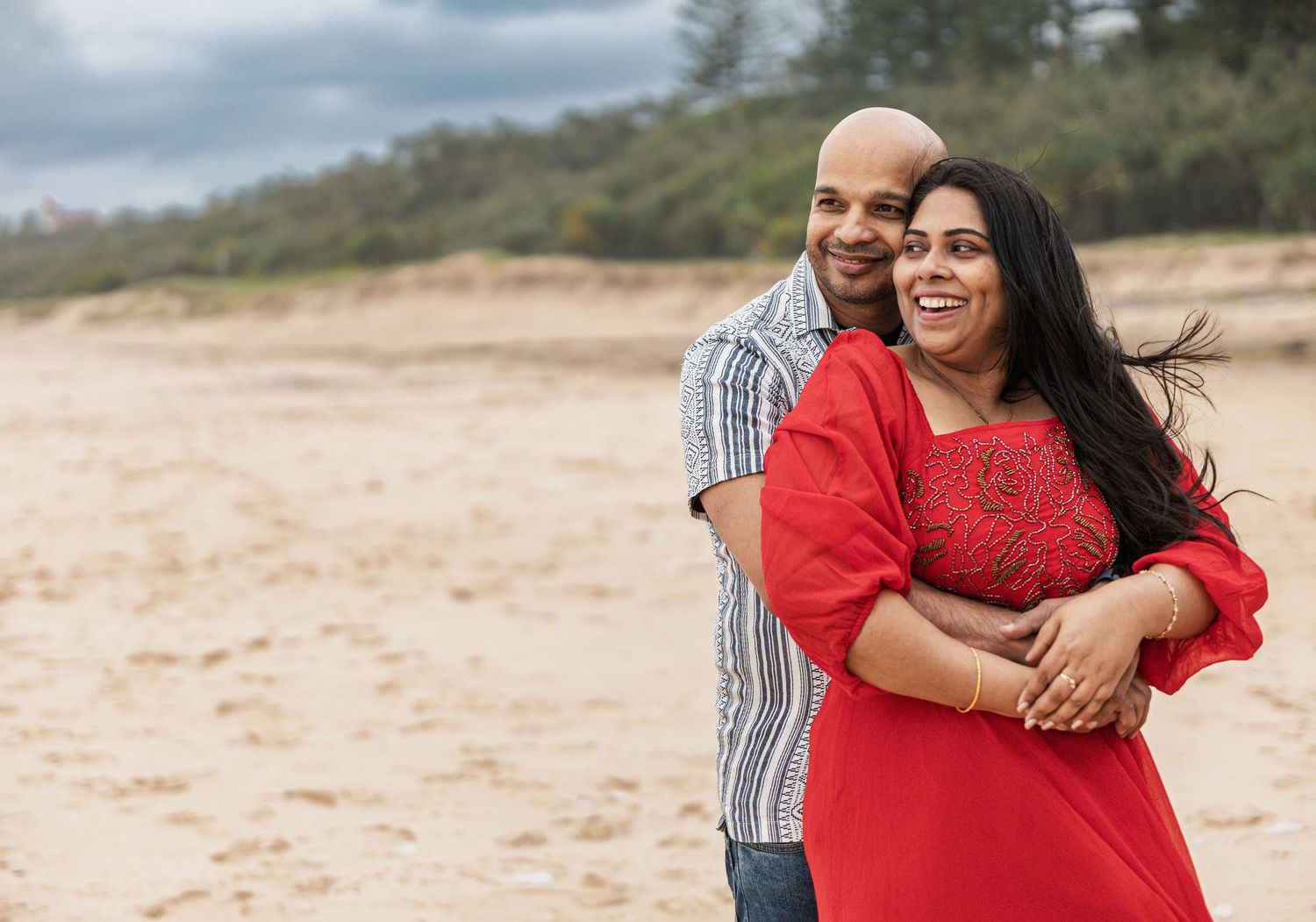 Couple embracing on beach with red dress and striped shirt against cloudy sky.