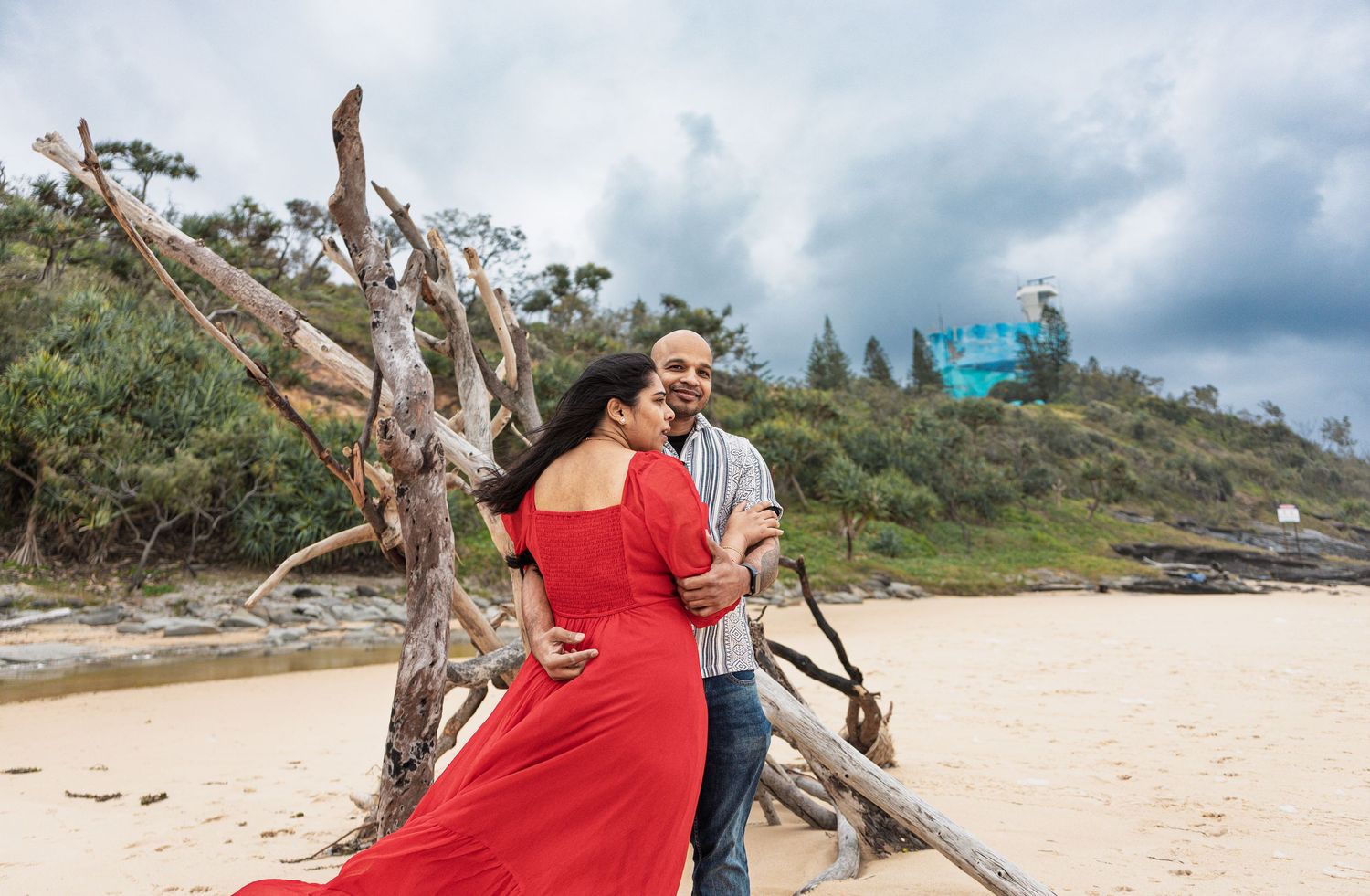 Couple walking on a scenic beach with driftwood and cloudy skies in the background.