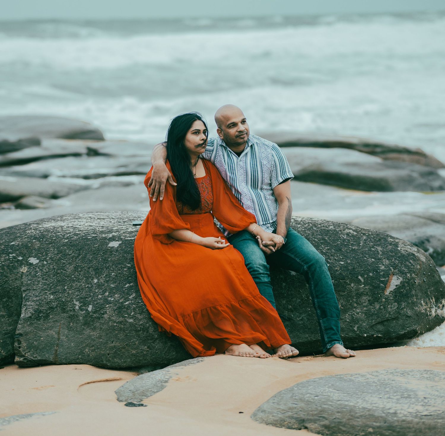 Couple in vibrant outfits pose romantically on rocky coastal formation by the sea.