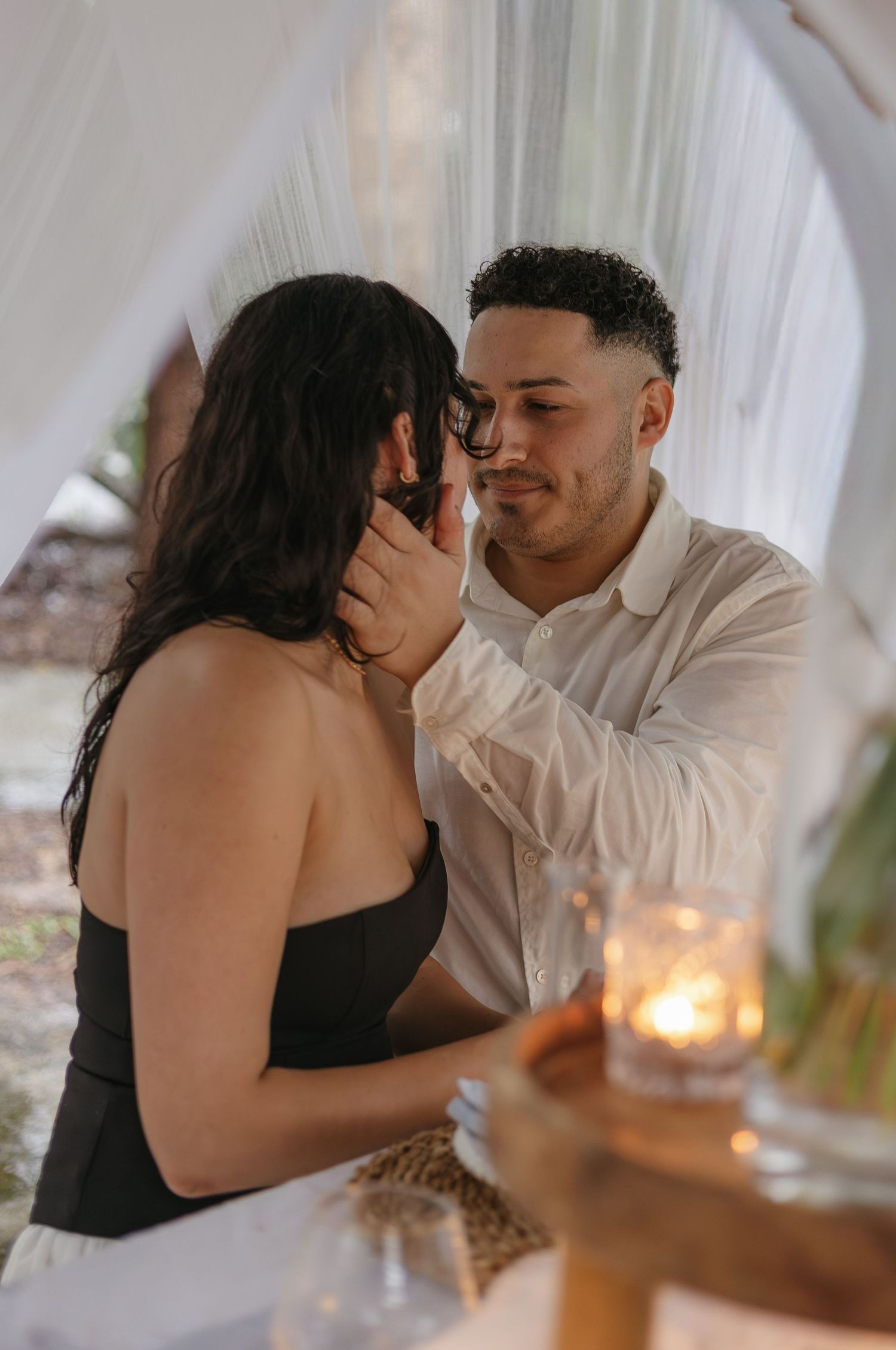 Couple embracing in romantic setting with white drapes and candlelight.