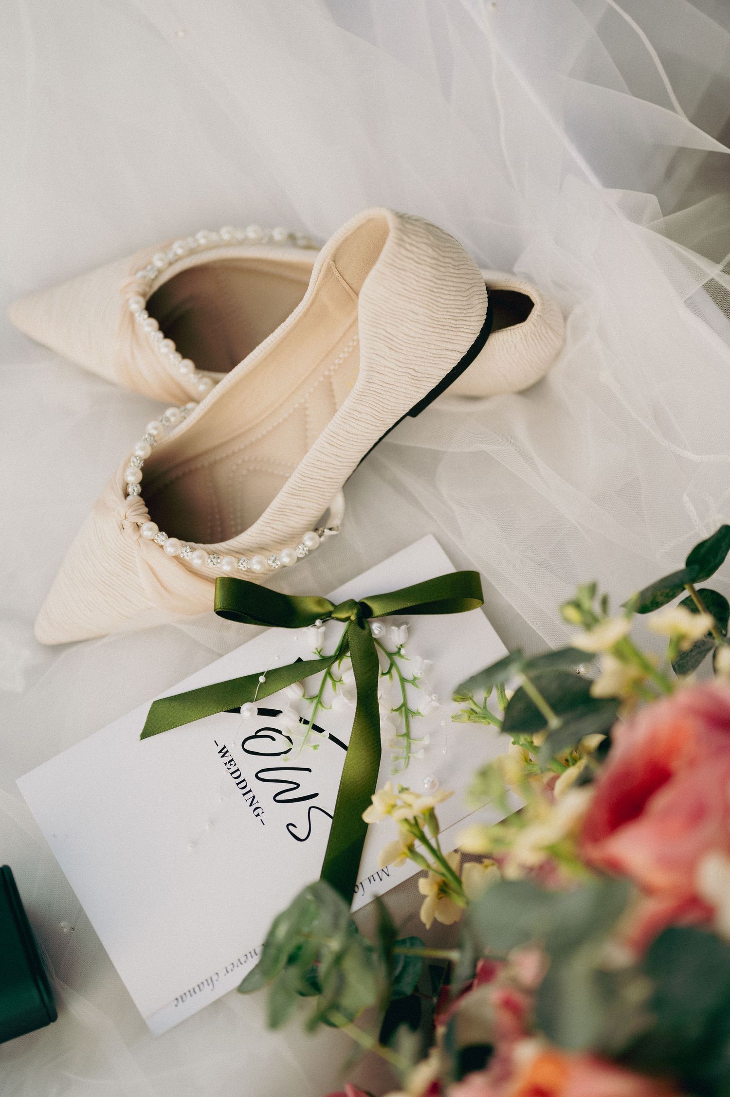 Elegant cream wedding shoes with pearl details and green ribbon tags on white tulle background.