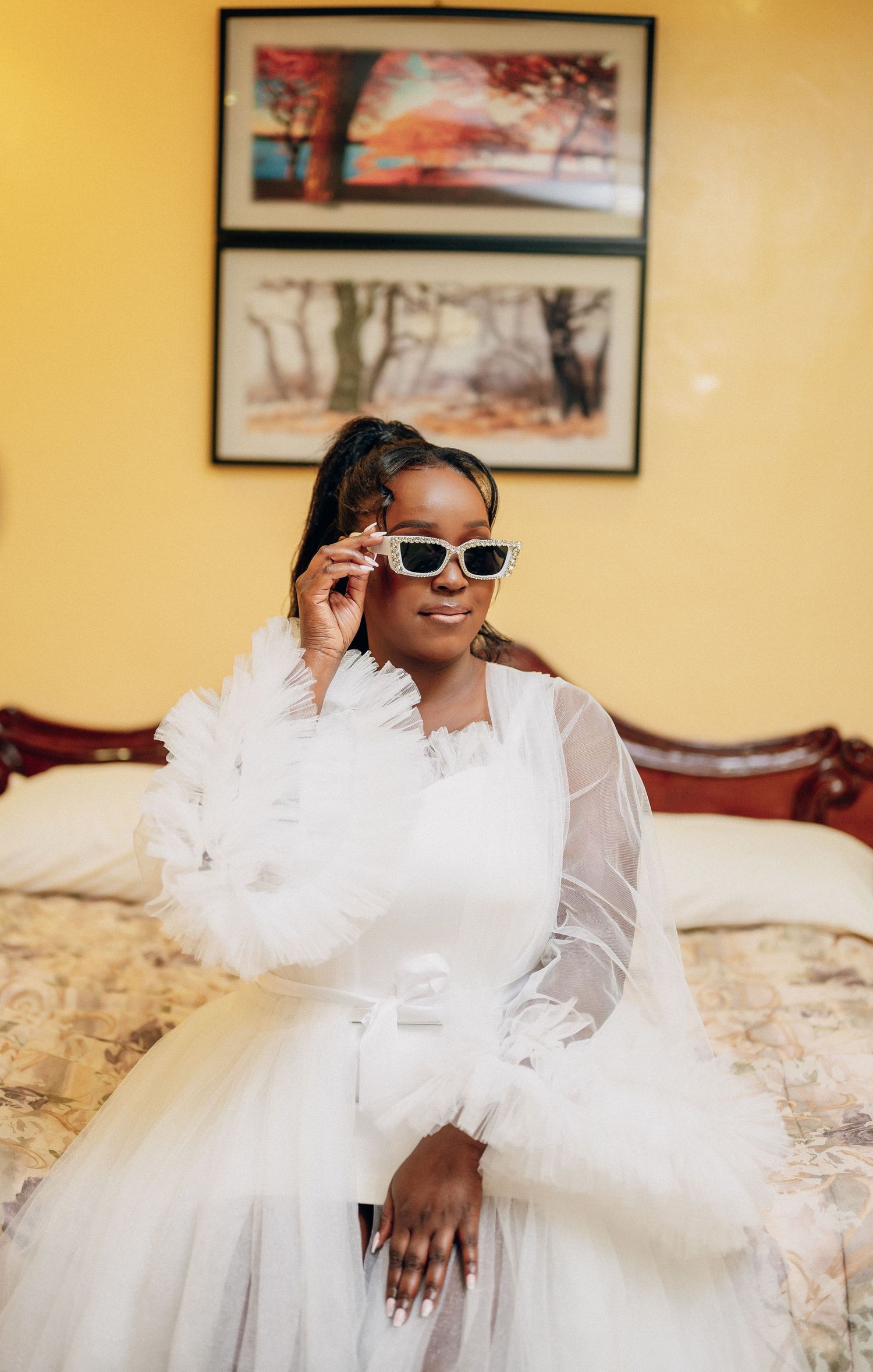 Person in flowing white tulle dress sitting on ornate furniture against yellow wall with framed artwork.