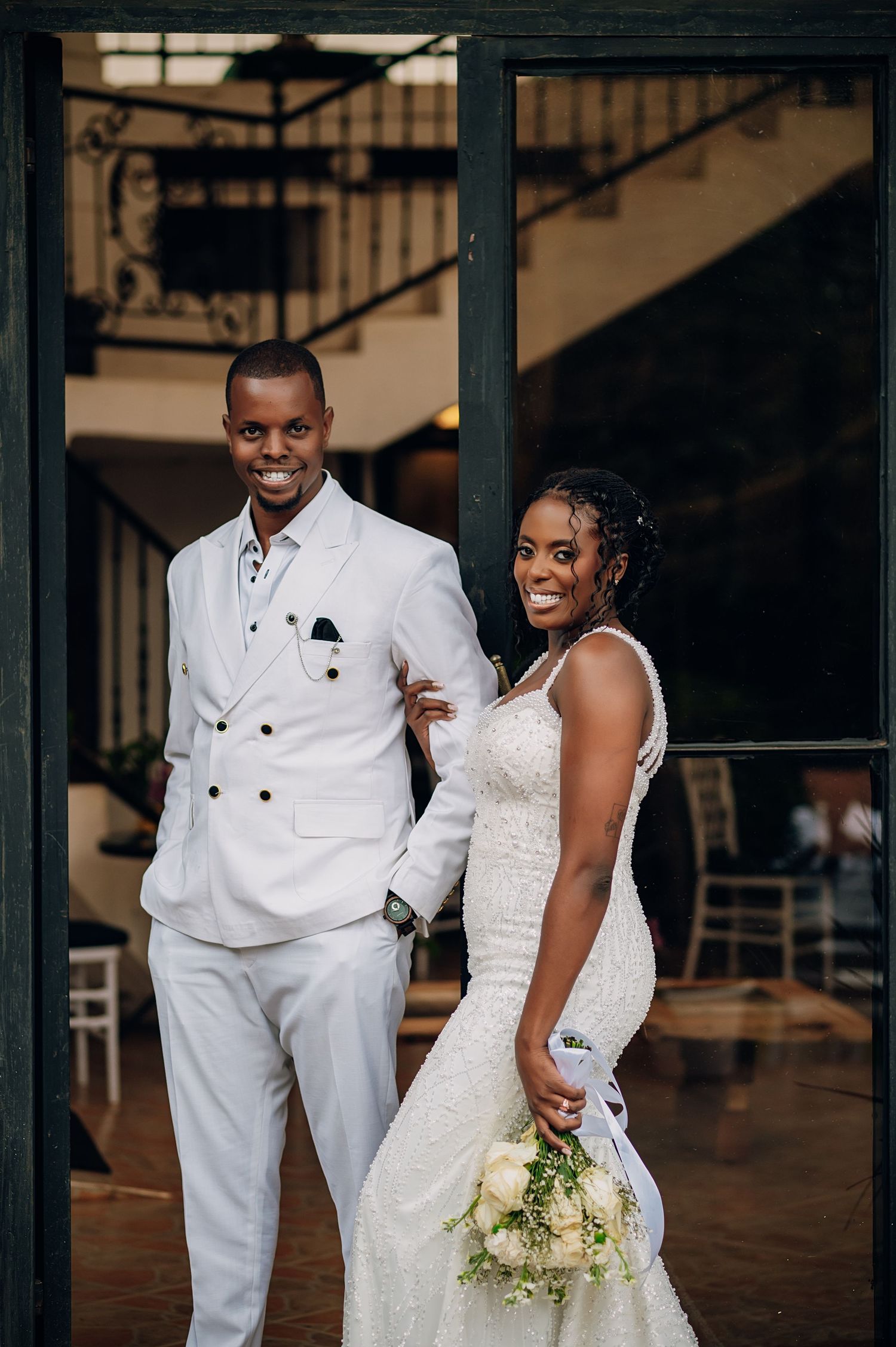 Wedding couple in white attire pose elegantly in front of modern architectural backdrop.