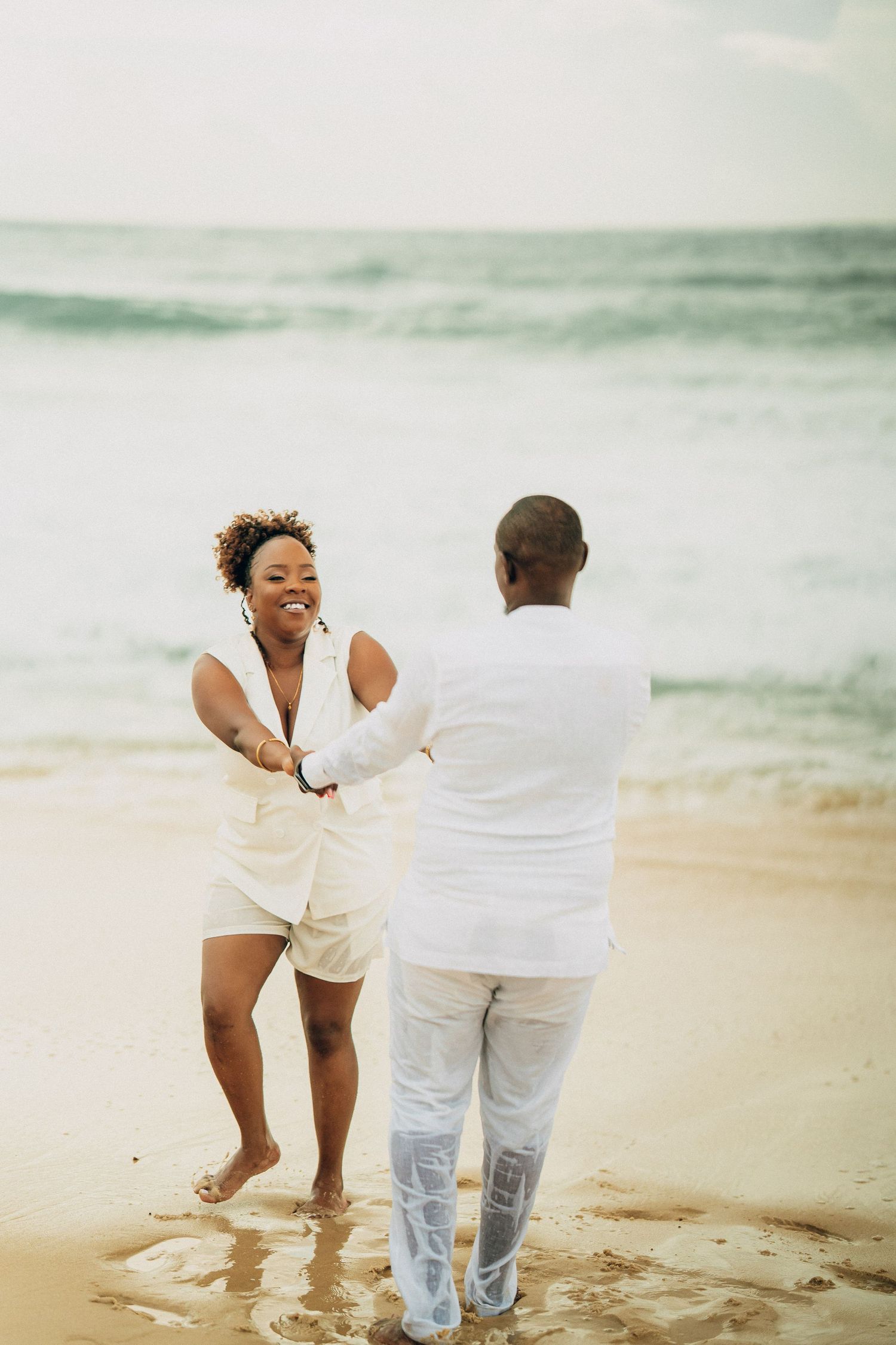 A couple shares playful moments on a sandy beach wearing white outfits.