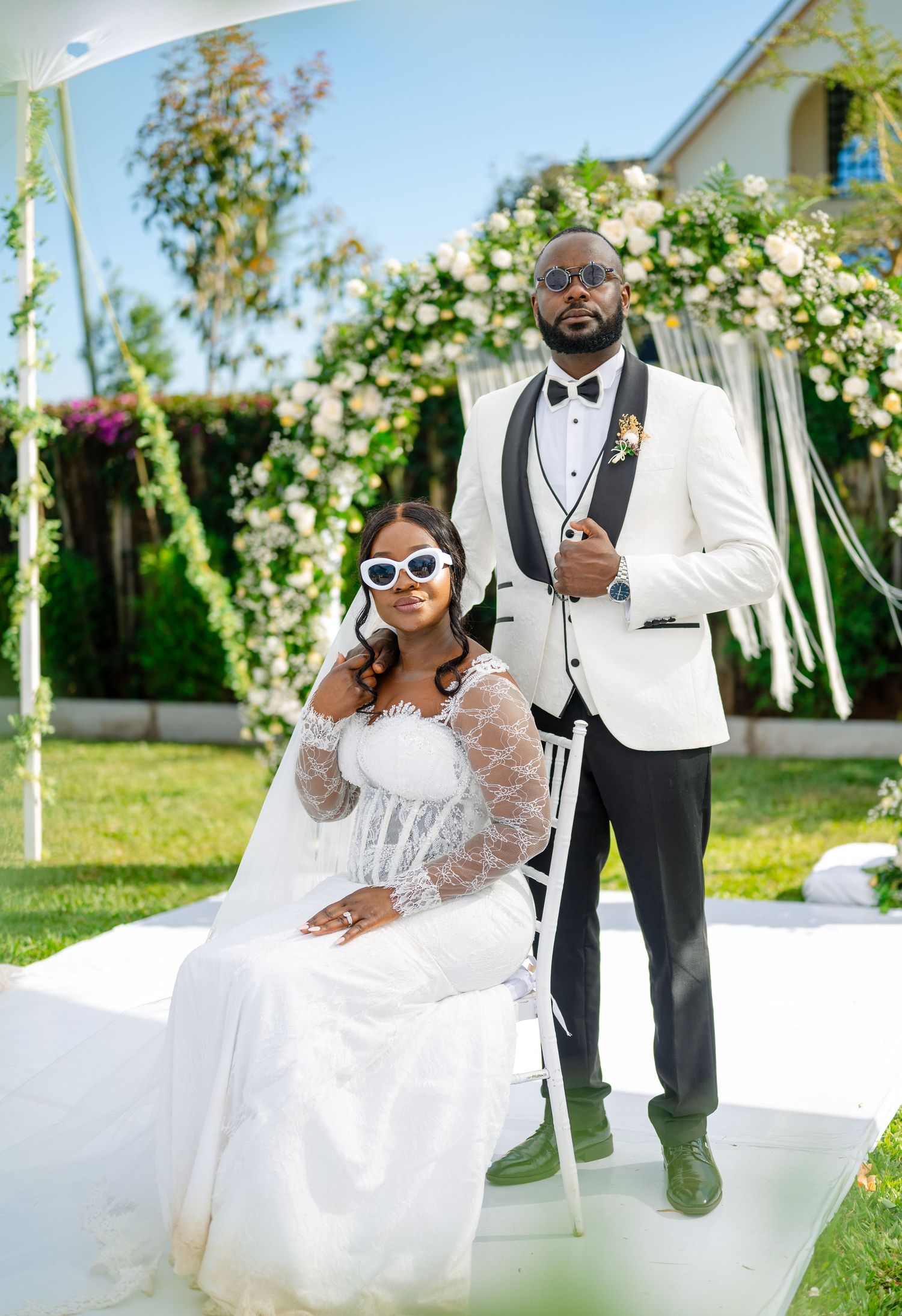 Stylish wedding couple in white attire pose under floral arch with sunglasses.