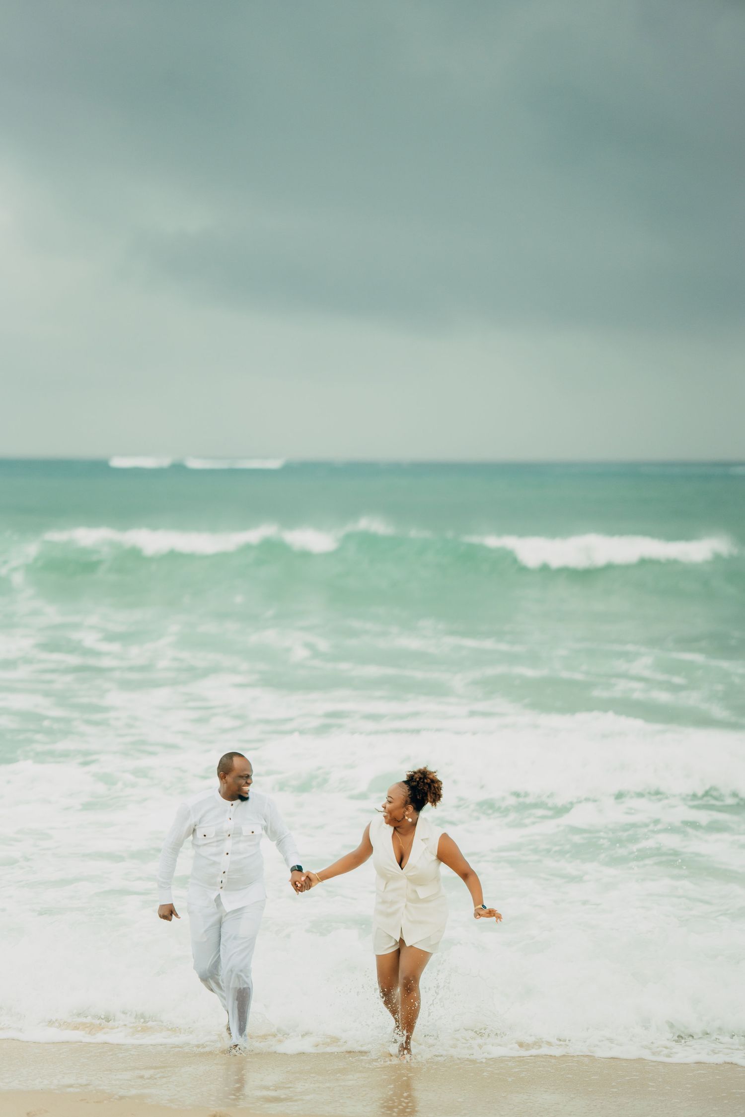 A couple in white clothing playing and dancing on a beach with turquoise waves in the background.