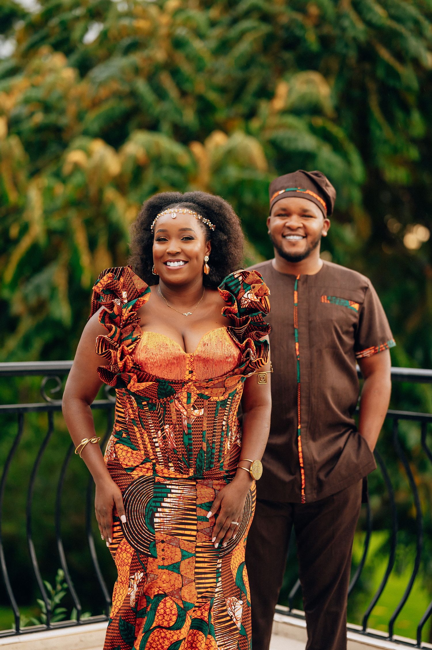 Couple wearing vibrant African print outfits pose together on a balcony.