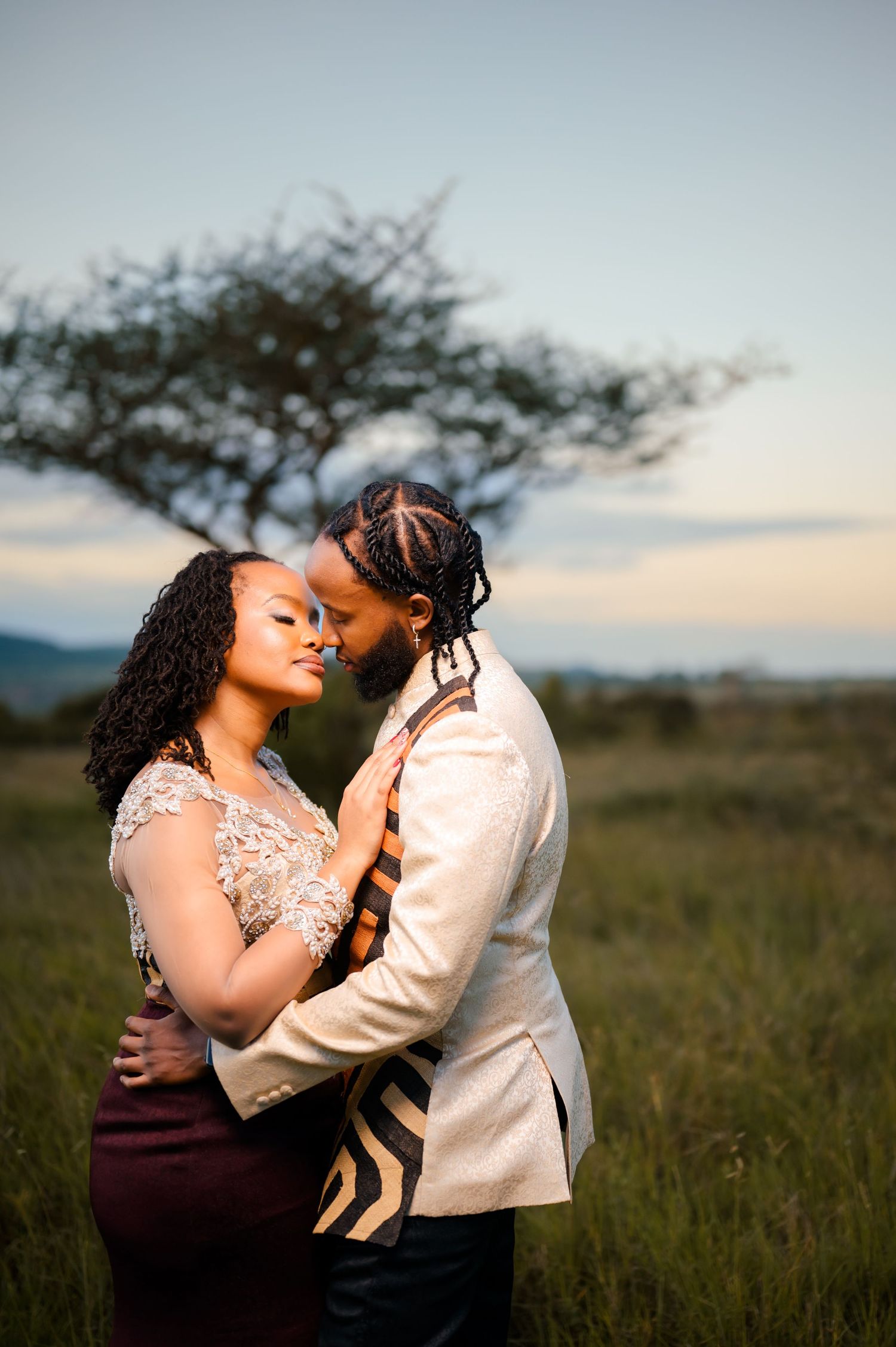 Couple embracing in a natural field setting with sunset sky in background.