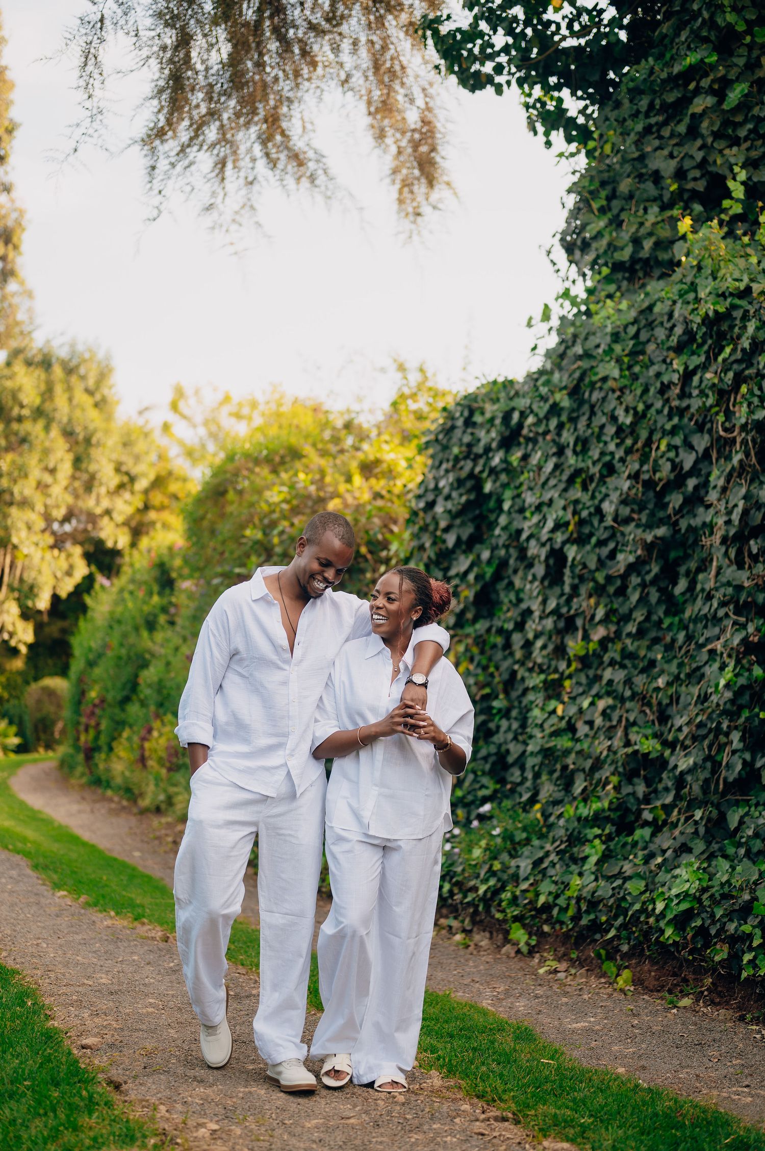 A couple in matching white outfits walking along a garden path lined with green foliage.