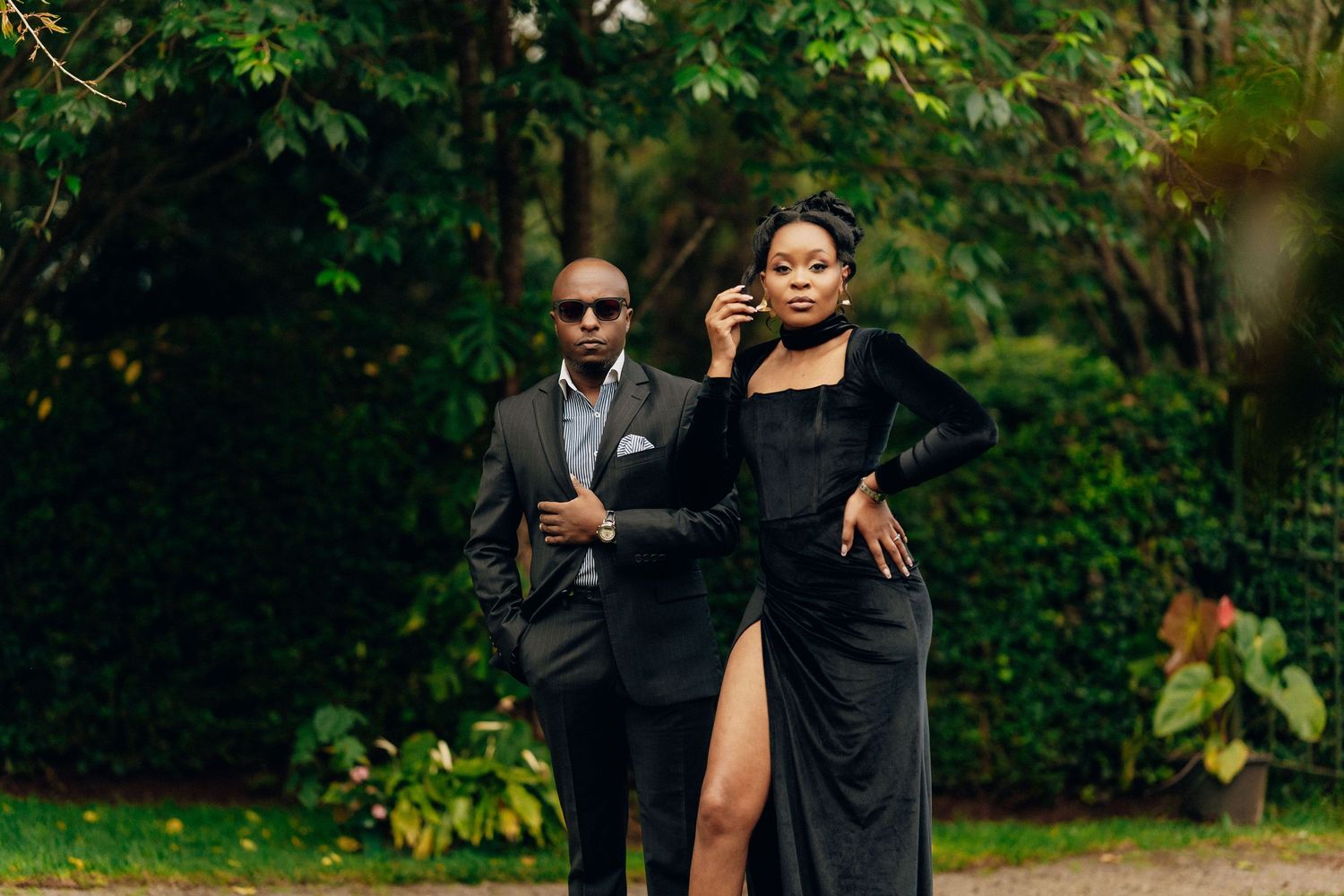 Glamorous couple poses in formal black attire against lush garden backdrop.