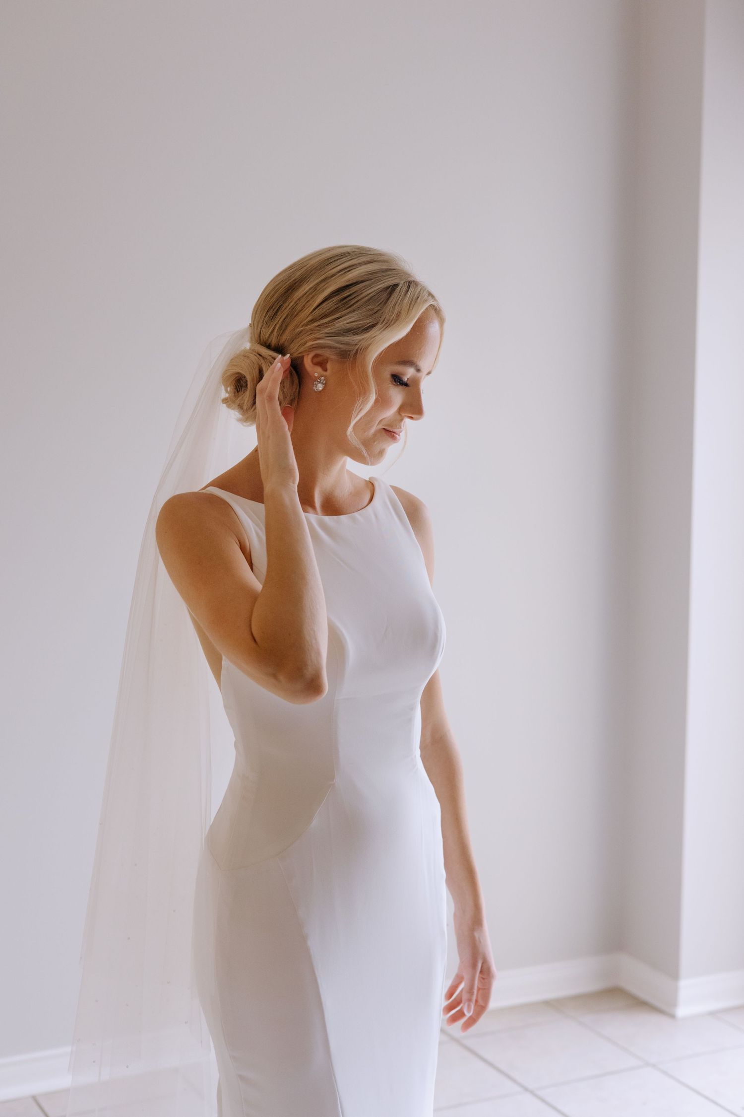 An elegant bride in a sleek white fitted gown with veil adjusts her earring in a minimalist bright room.