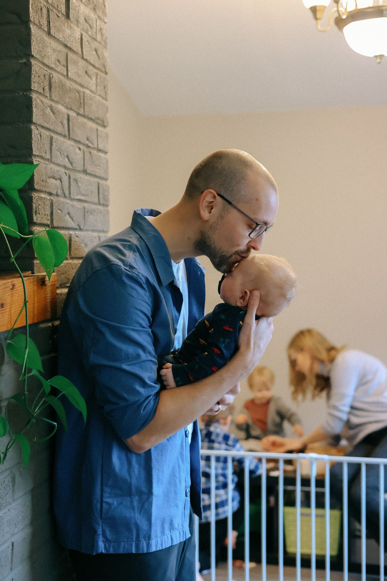 Bald bearded father gently kisses his sleeping newborn baby while standing by a brick wall.
