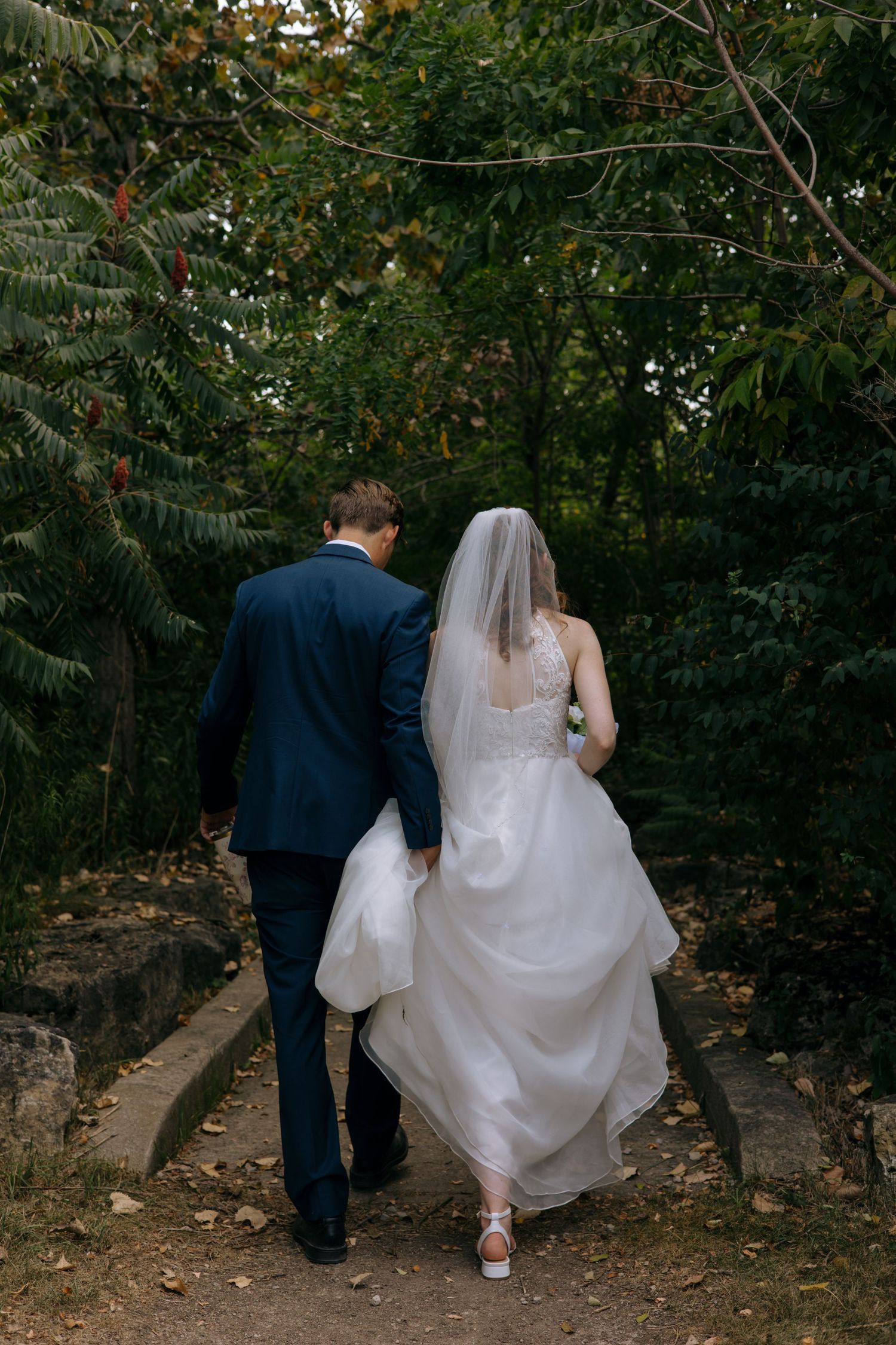 Bride and groom walking hand in hand through a lush green forest path after wedding ceremony.