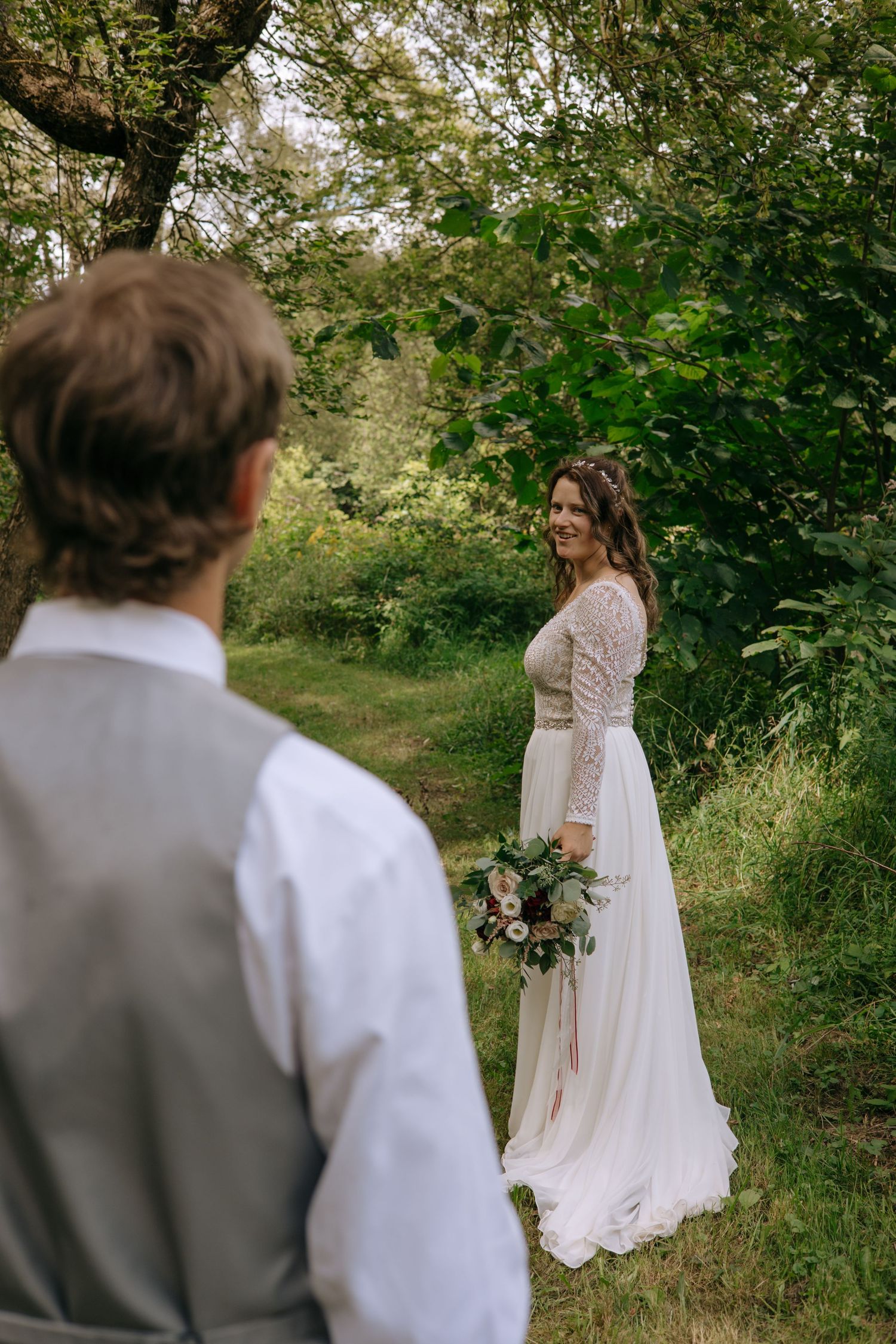 Groom watches as smiling bride in lace wedding dress holds bouquet on a lush green forest path.