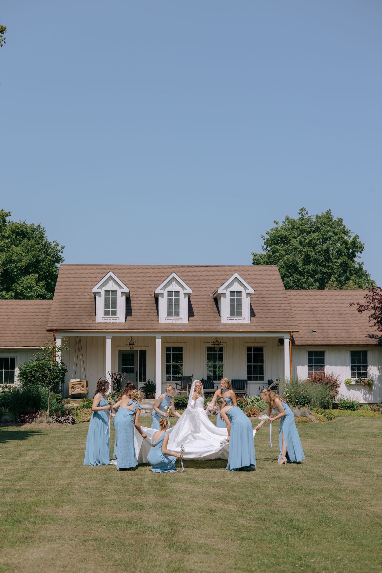 Bride in white gown with bridesmaids in light blue dresses posing on green lawn in front of charming white farmhouse.