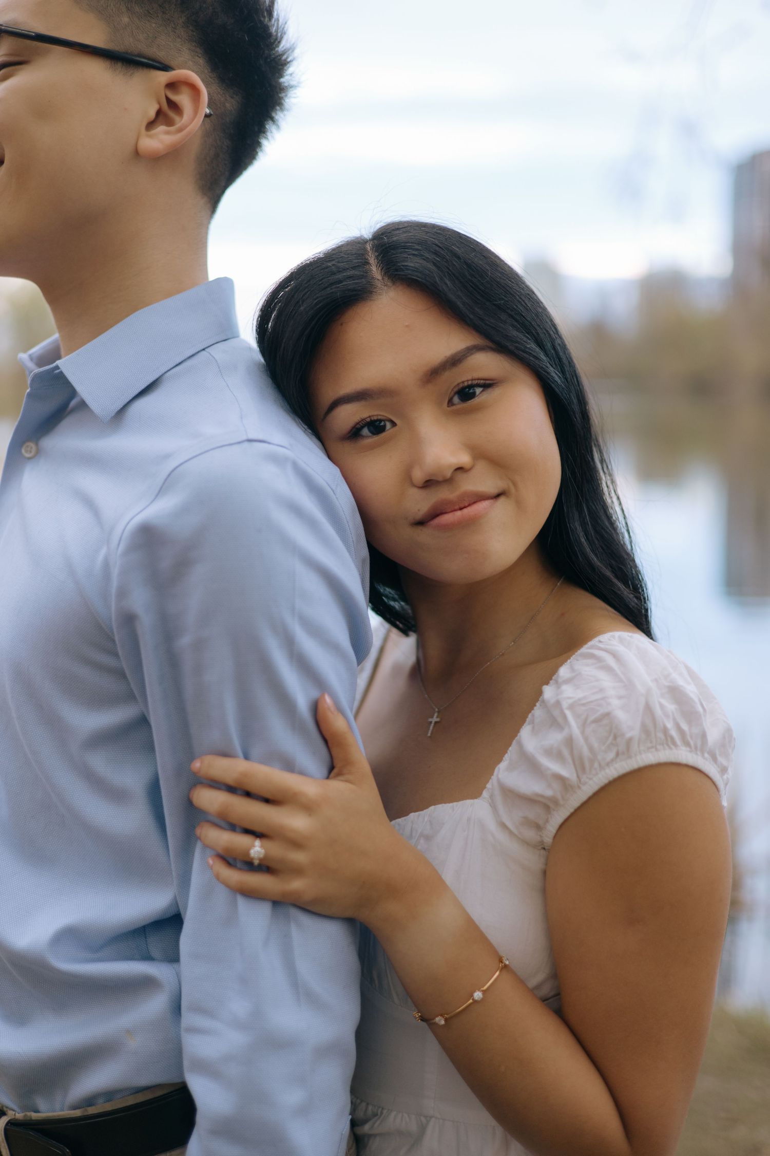 Young Asian woman in white dress smiling at camera while leaning against her partners arm outdoors.