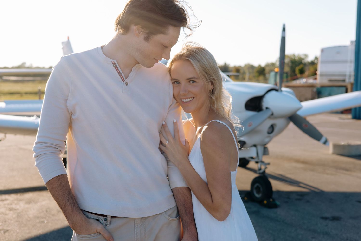 Smiling blonde woman in white dress leans against a man in a white shirt with a small aircraft behind them.
