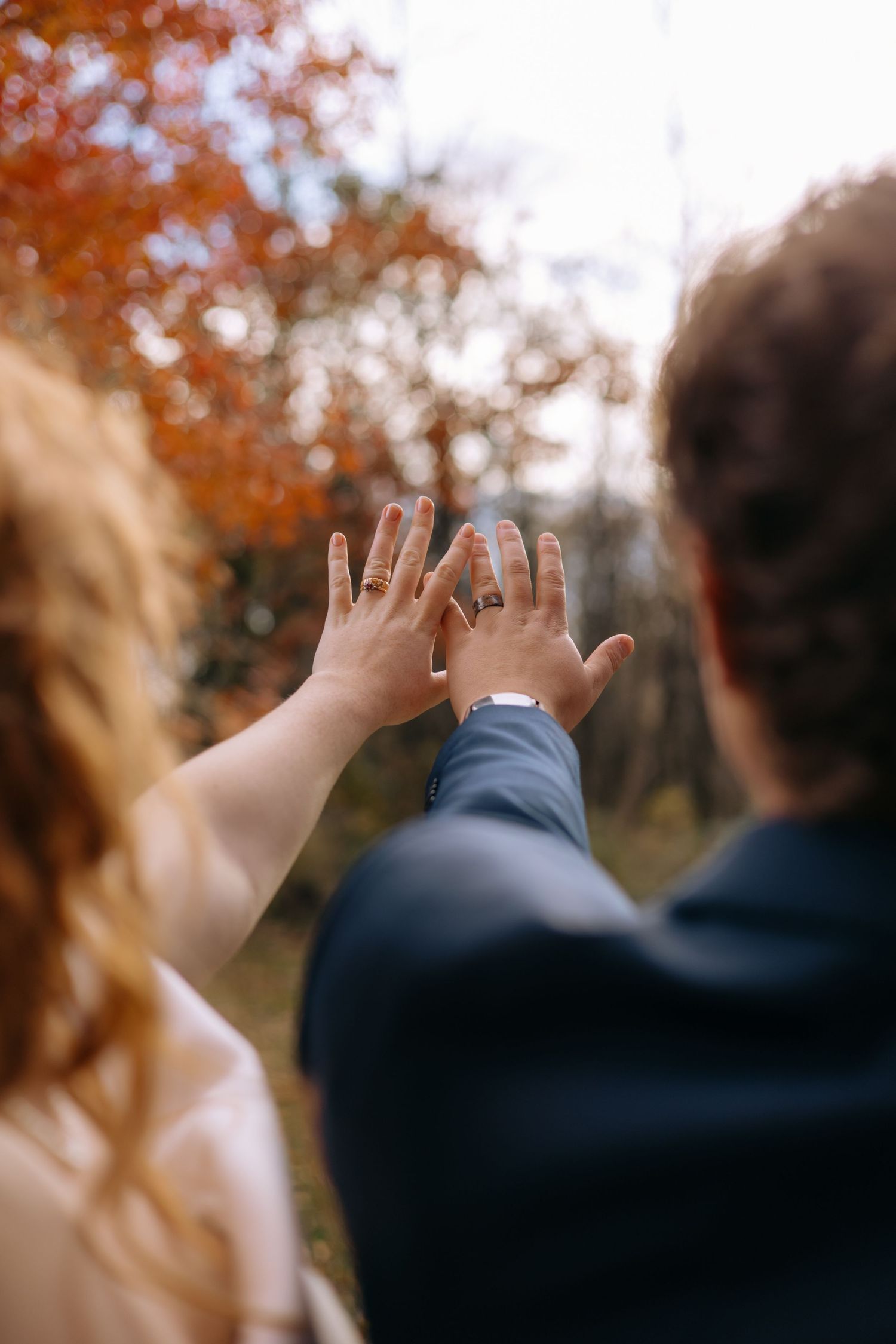Newlyweds hold up their hands displaying wedding rings against a colorful autumn forest backdrop.
