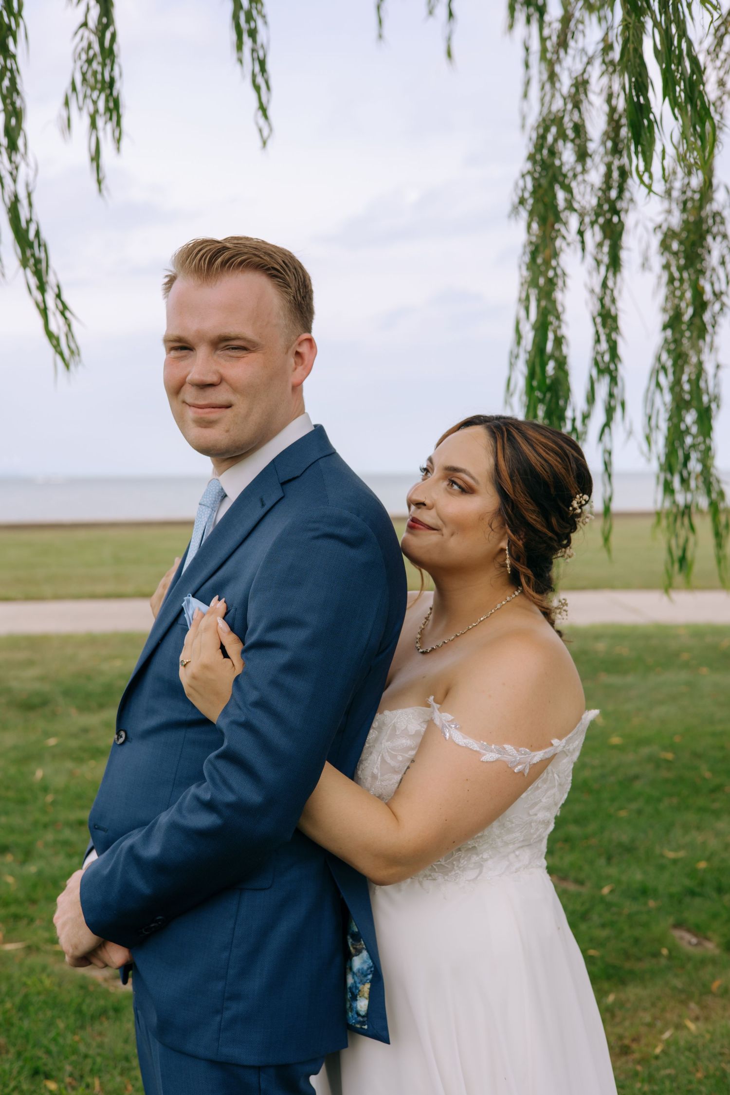 Bride in white lace gown embracing groom in navy suit near a weeping willow by the waterfront.