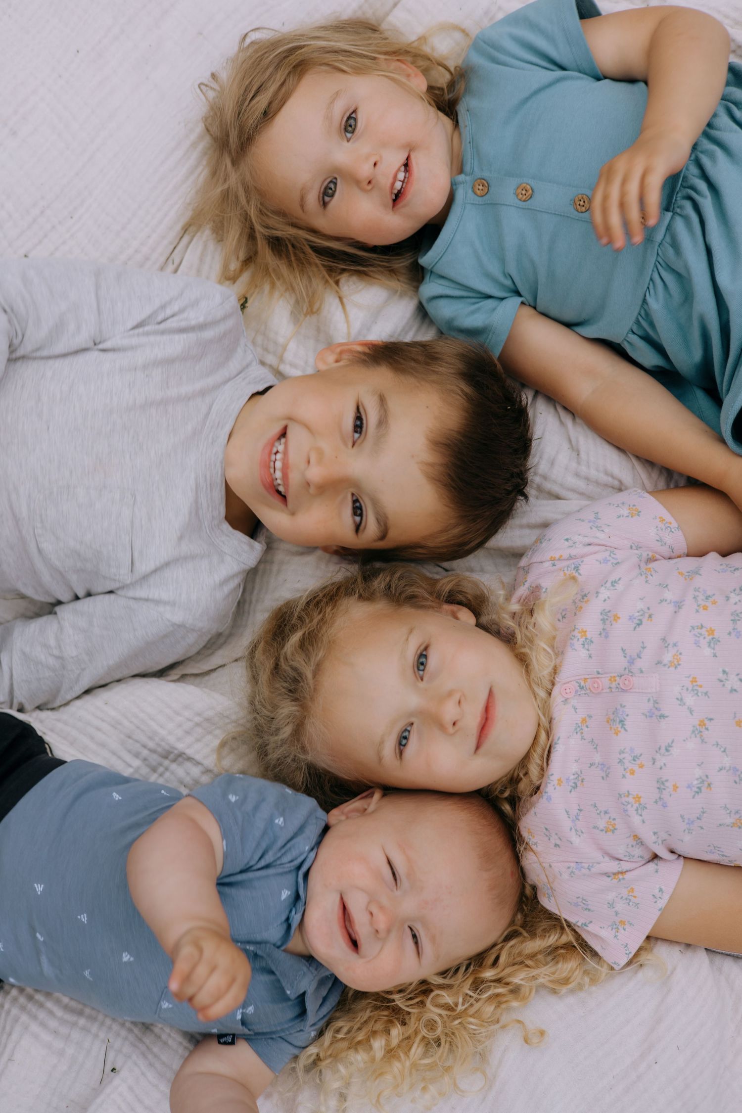 Four young siblings lying together on white bedding, smiling and laughing in a sweet overhead portrait photo.