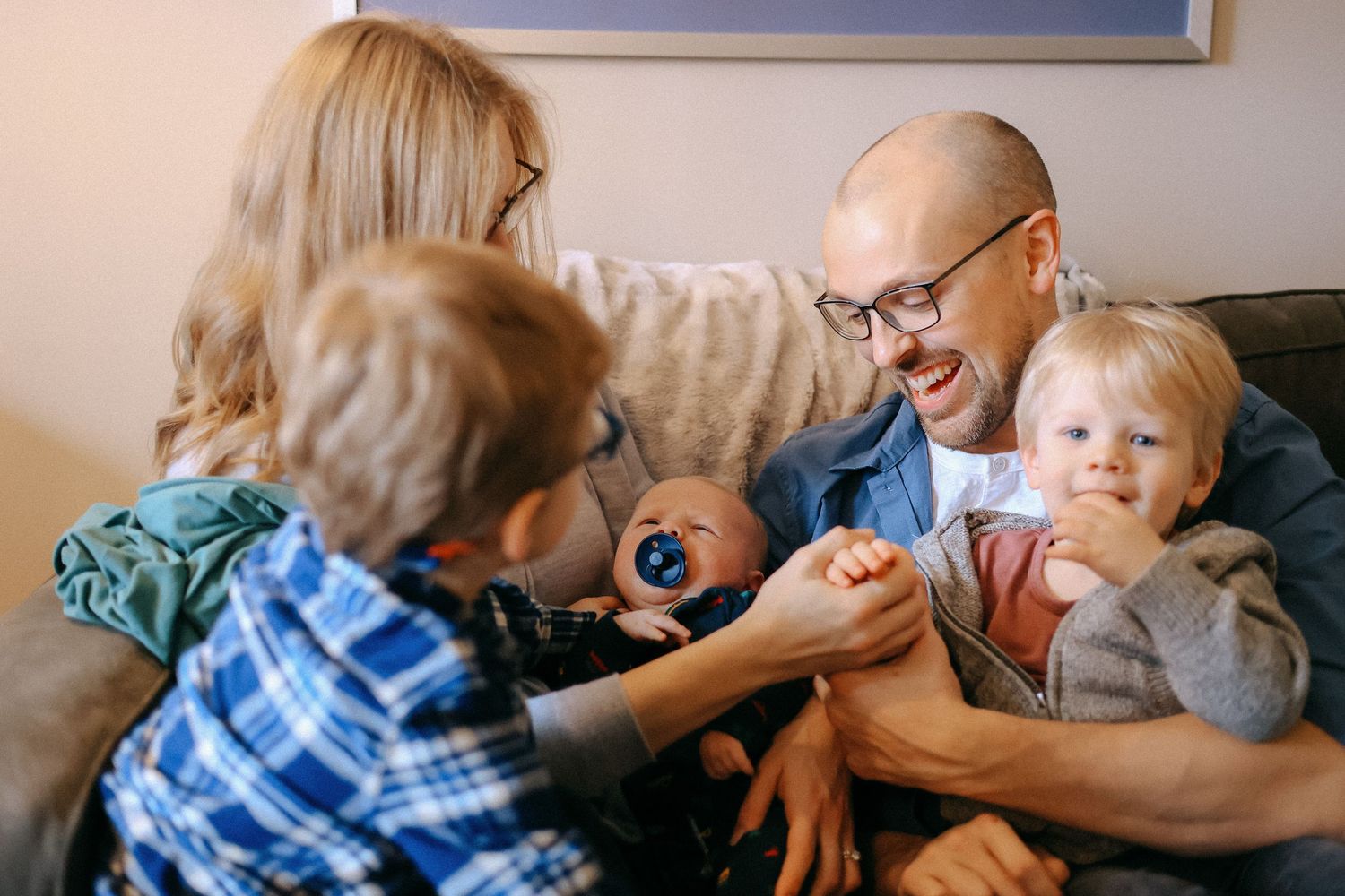 Family of five with newborn baby and two young children laughing together on sofa.
