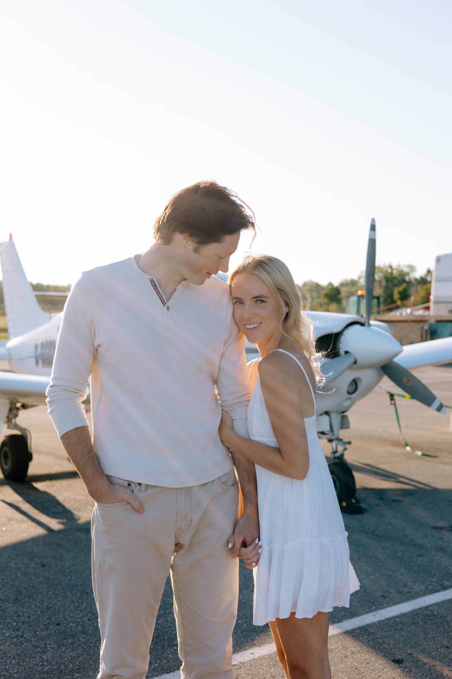 Couple posing together at an airport tarmac with a small propeller plane in the background during golden hour.