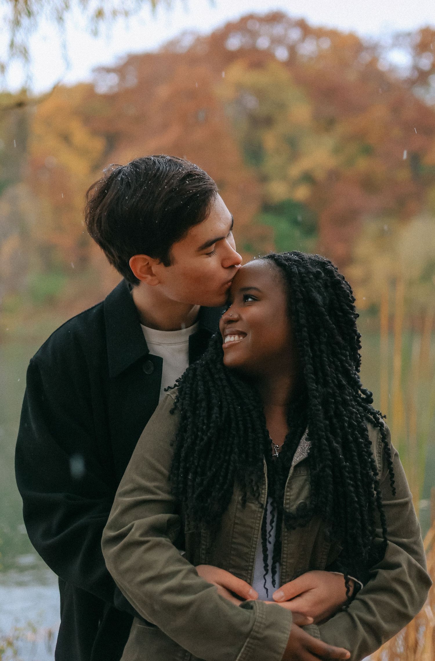 Man kisses woman on the forehead as she smiles near a lake surrounded by colorful autumn foliage.