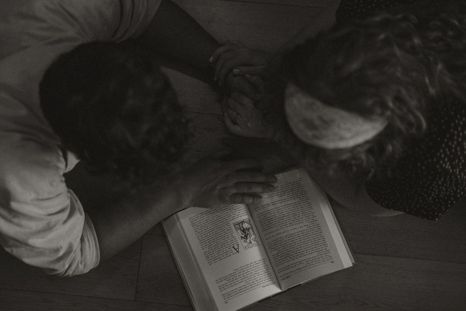 Black and white overhead view of couple reading an open book together on the floor.