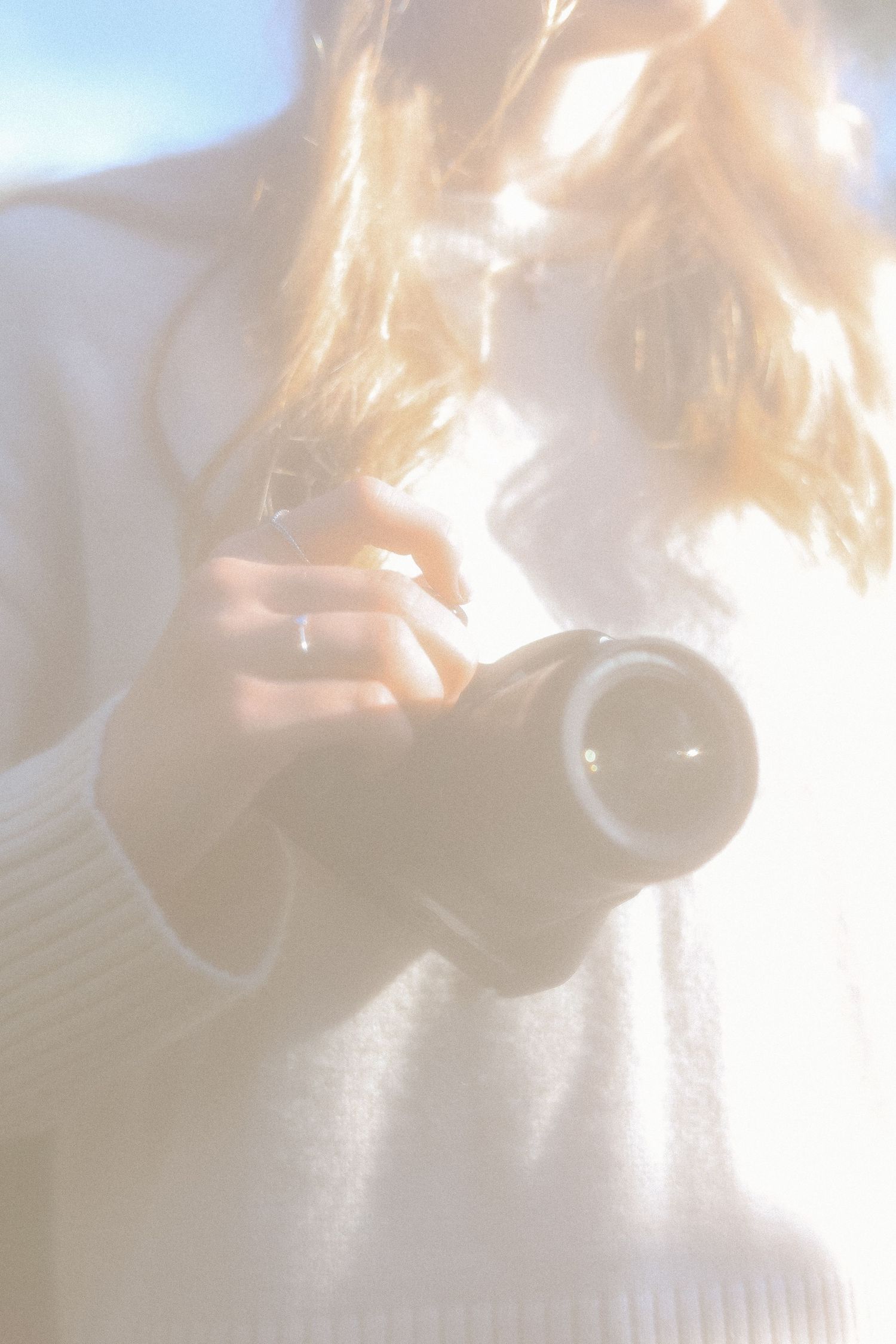 Softly lit photo of a blonde woman in white holding a camera lens outdoors in bright sunlight.