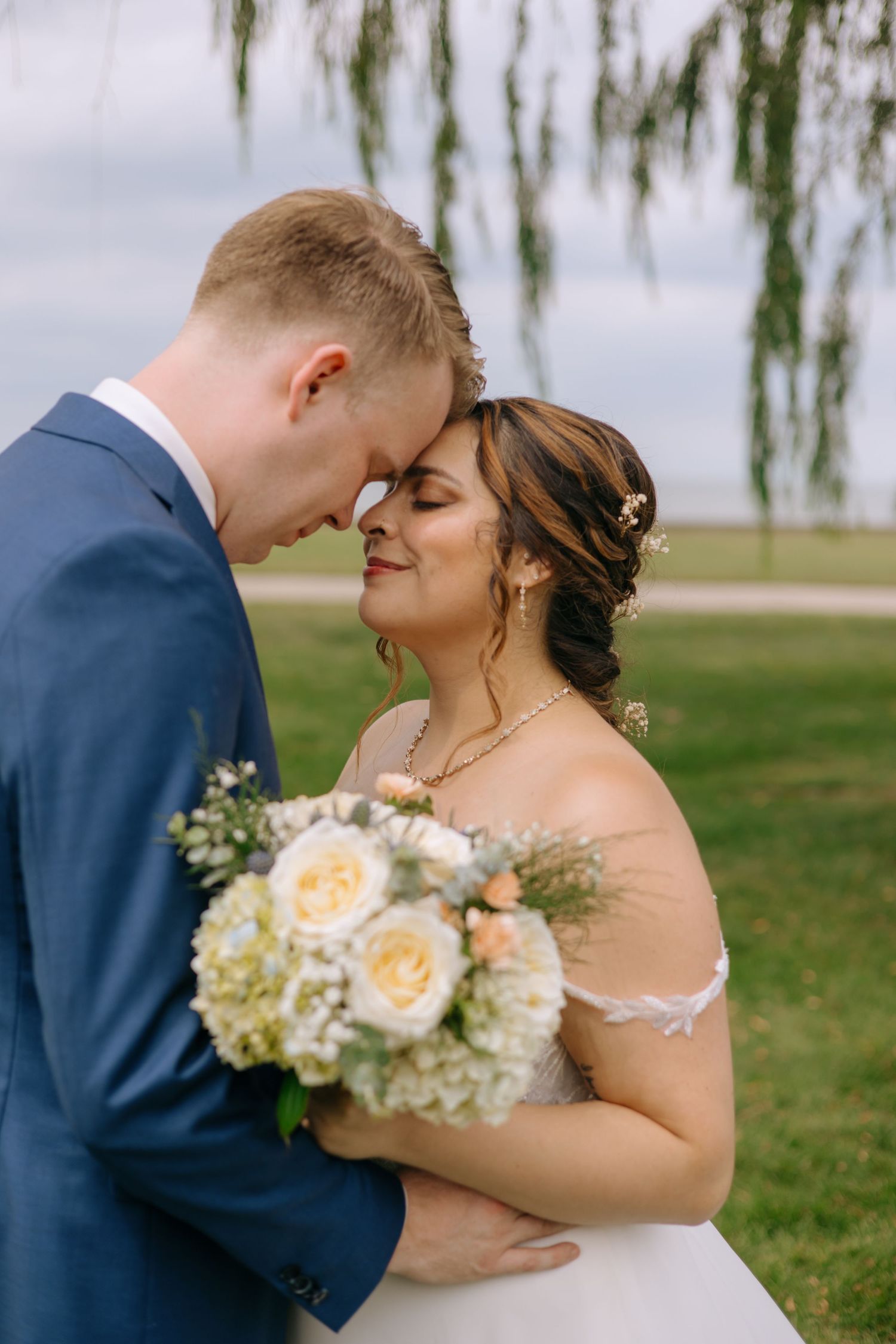 Bride and groom share a romantic forehead touch outdoors, bride holding a white and peach bouquet in a lush green setting.